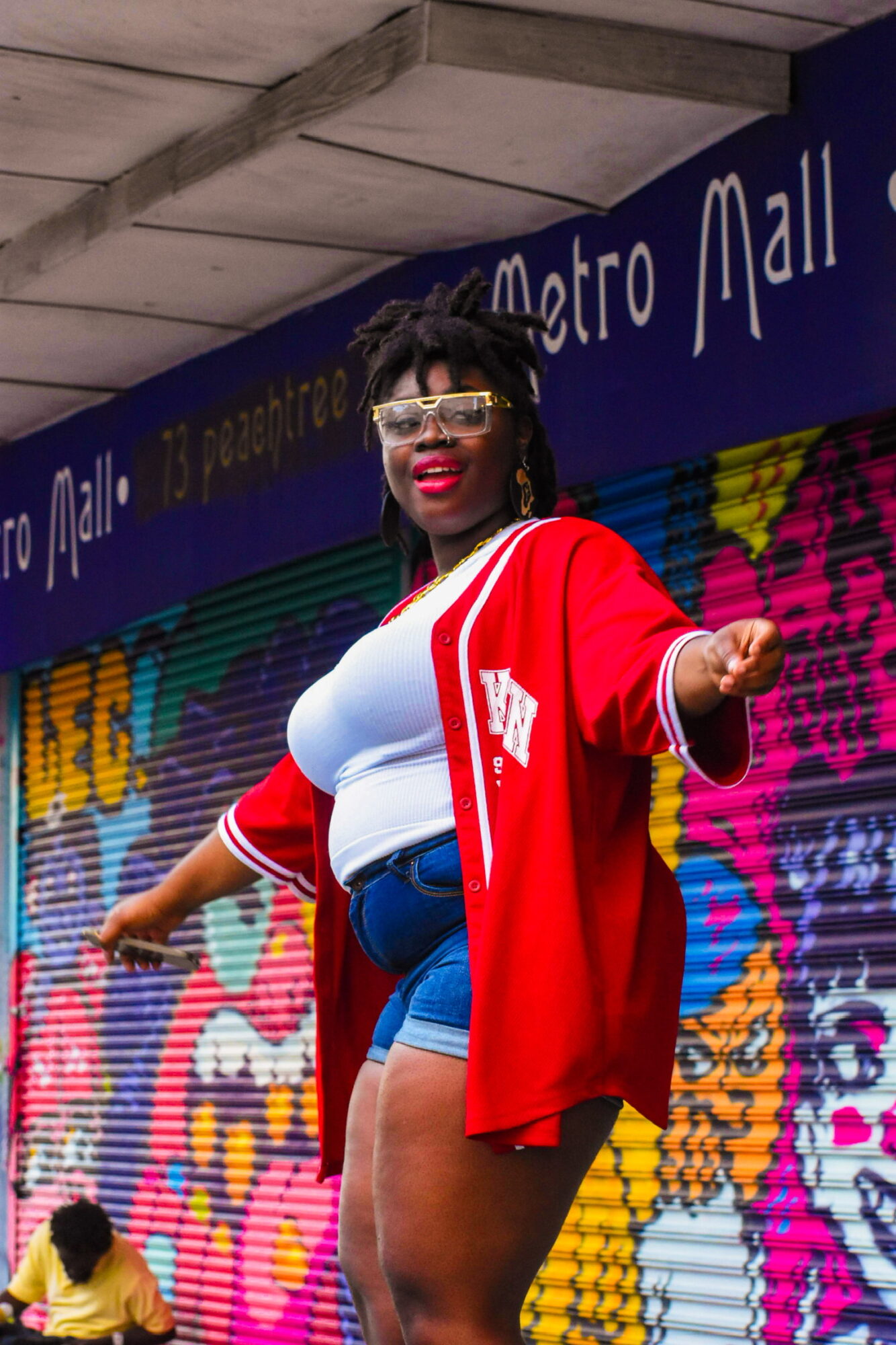 Woman with curly hair and glasses smiling, wearing a red jacket, white top, and denim shorts, standing in front of colorful mural at Metro Mall.