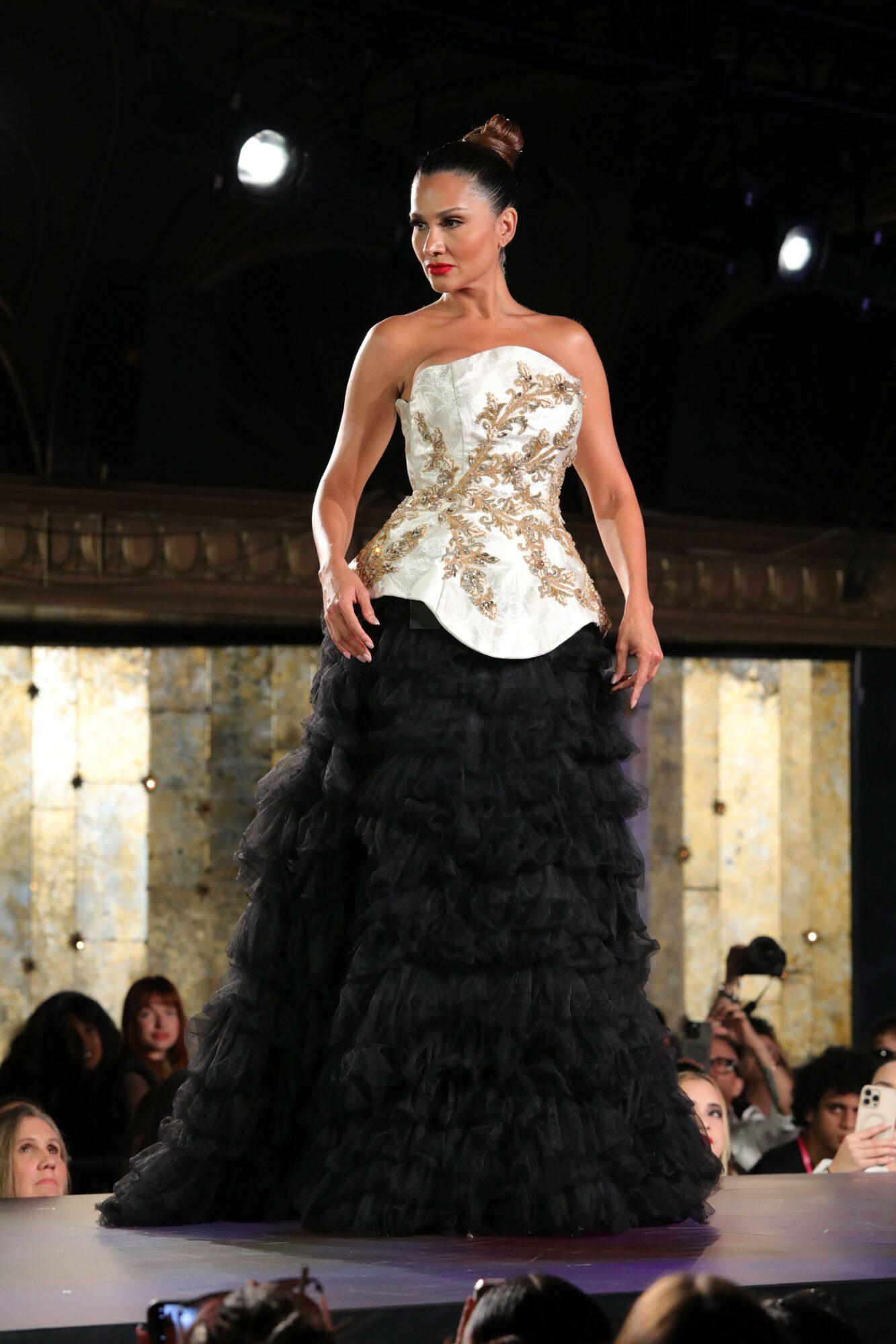 Woman in a white dress walking on a runway with audience watching, dark background, and wooden wall behind her.