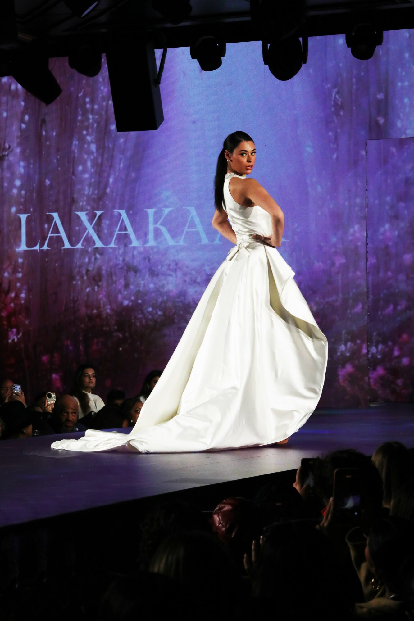 Model in a white gown on runway with purple background and 'LAXAKA' text, audience visible in foreground.