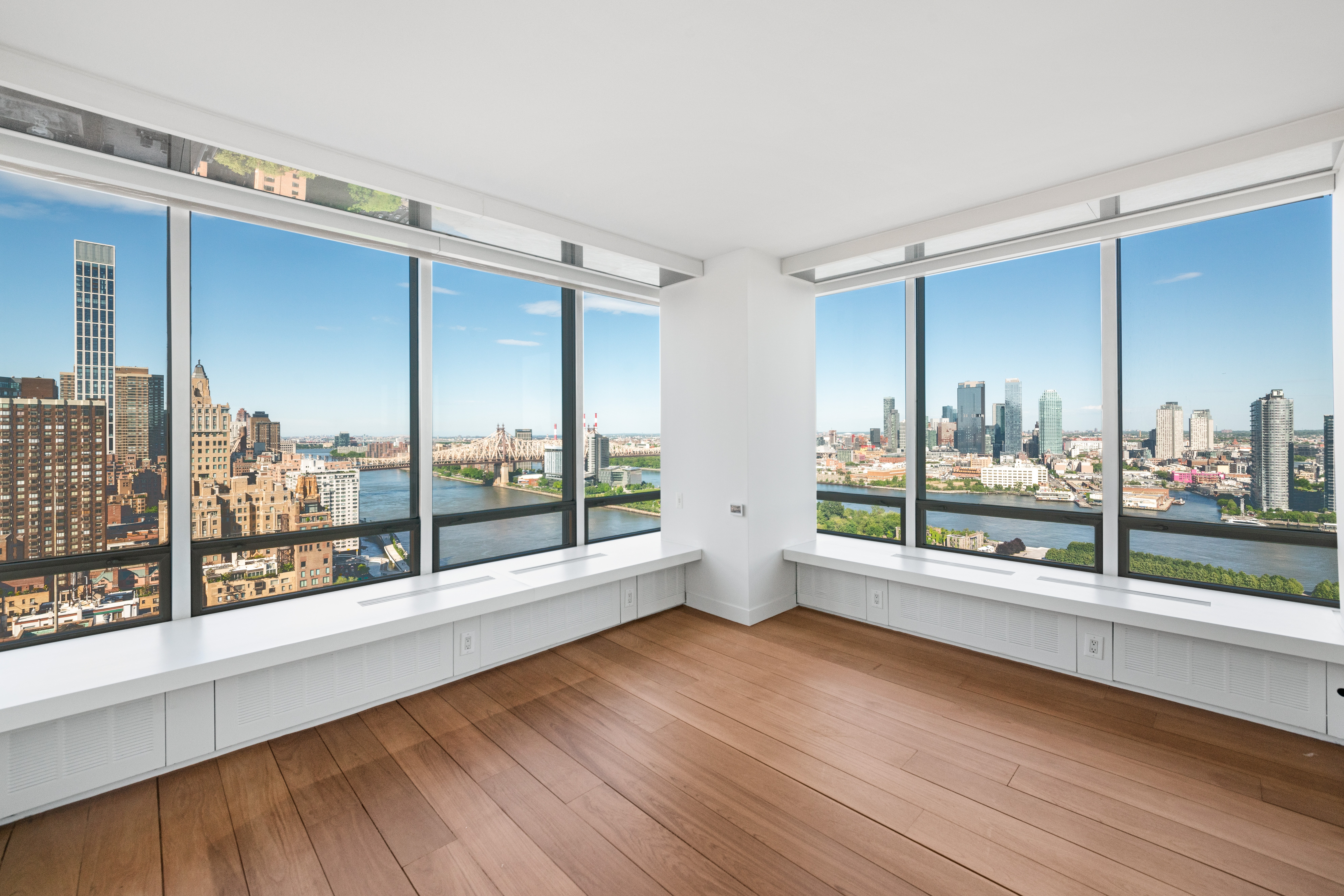 Empty room with large windows showing city skyline and river, wooden floor, white walls, and ceiling.