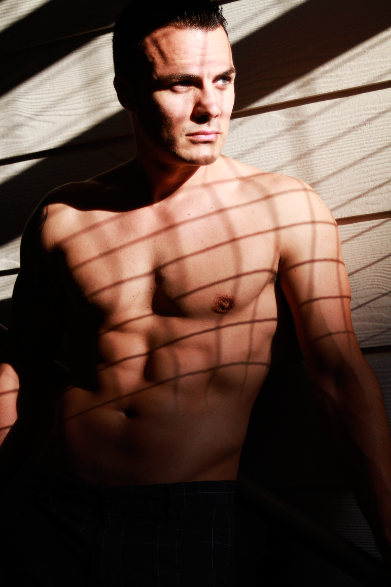 Man with short dark hair and bare chest, shadows from blinds on his face and body, looking to the side.