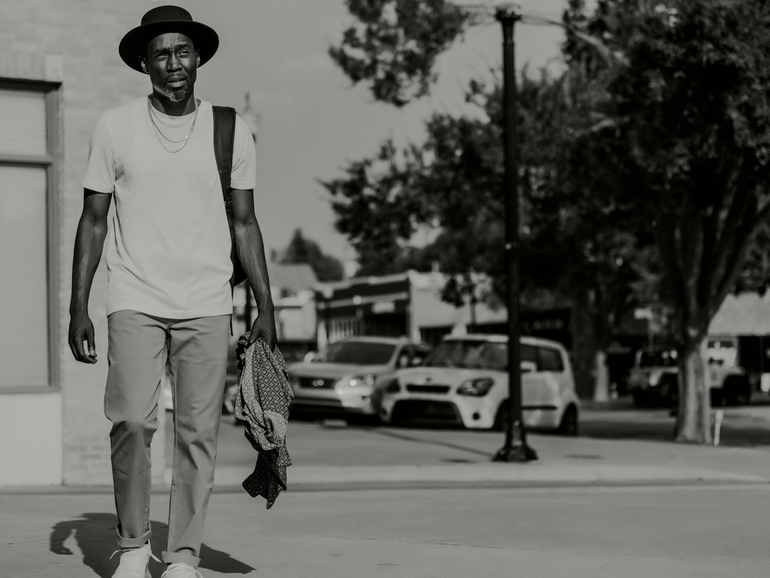 Man walking on sidewalk with trees and parked cars in background, wearing a hat, t-shirt, and pants.