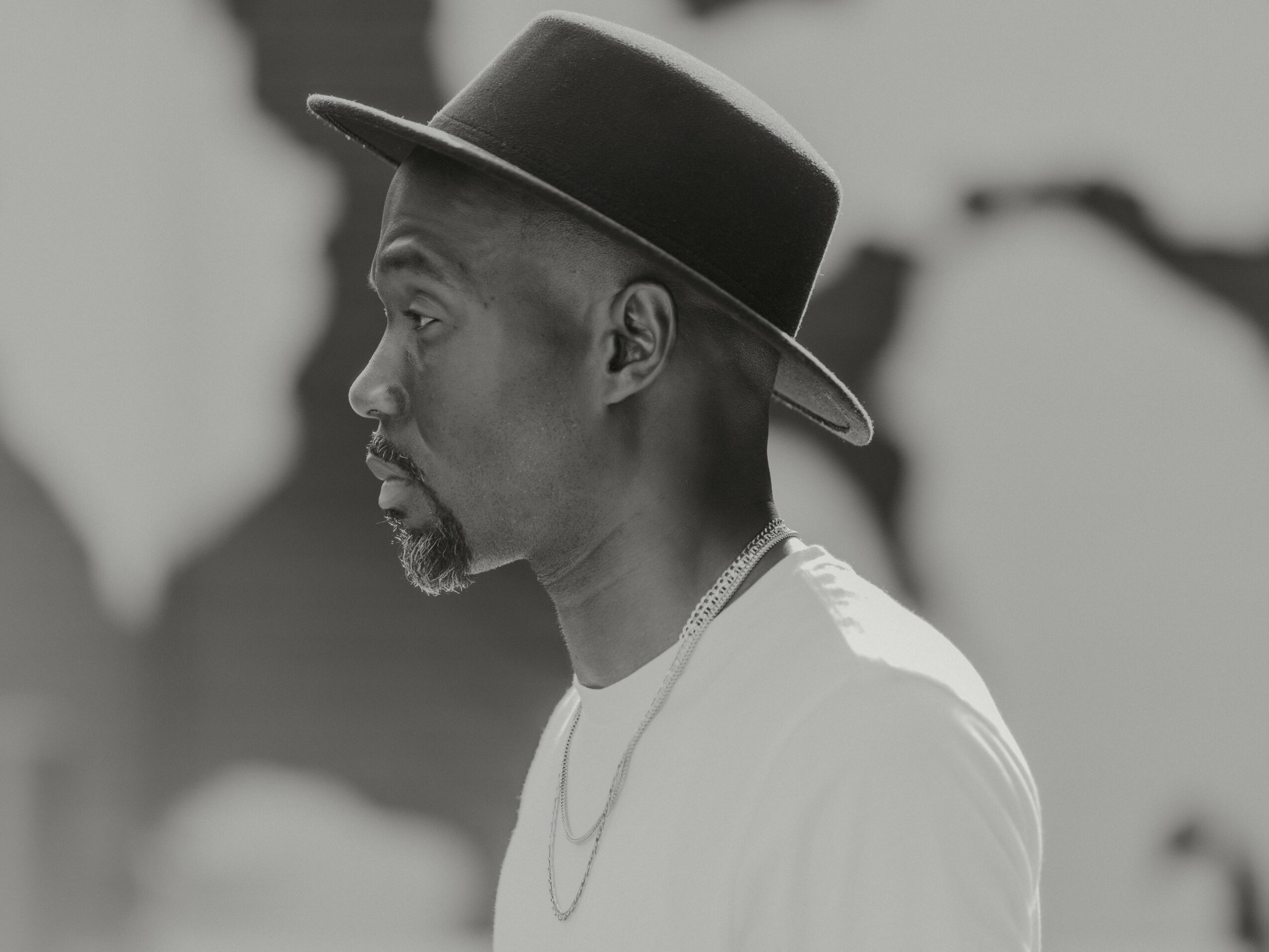 Side profile of a man wearing a hat and a necklace, in black and white.
