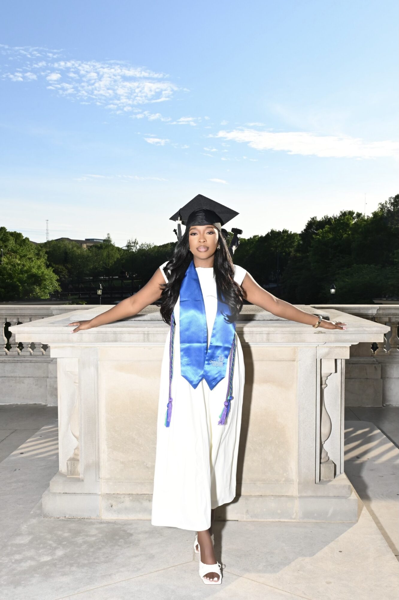 Graduate in cap and gown standing outdoors with arms outstretched on a stone structure, blue sky in background.