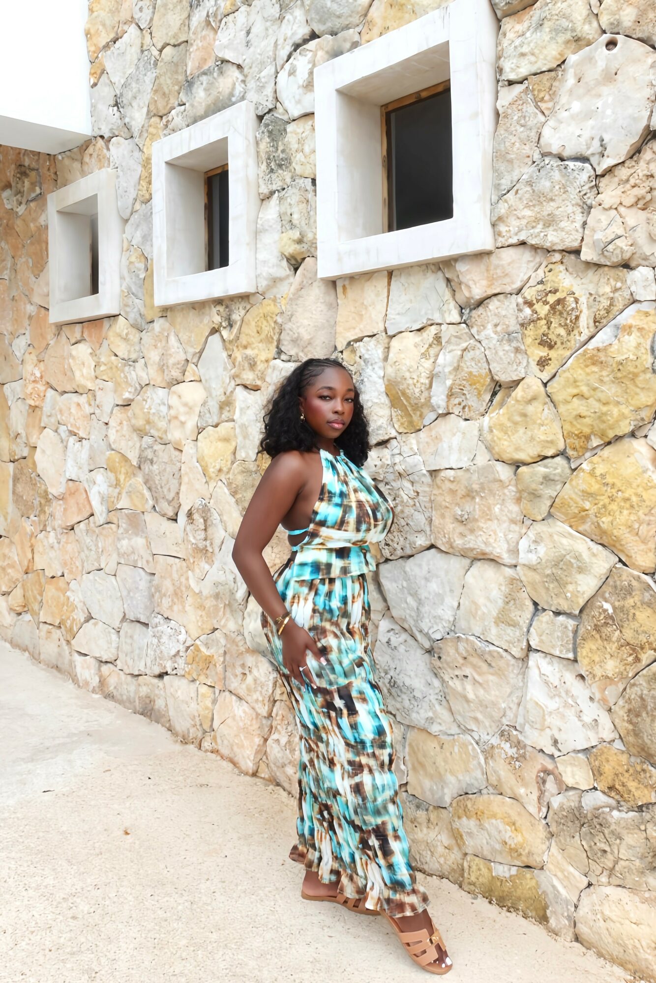 Woman in a colorful dress standing against a stone wall with three small square windows above.