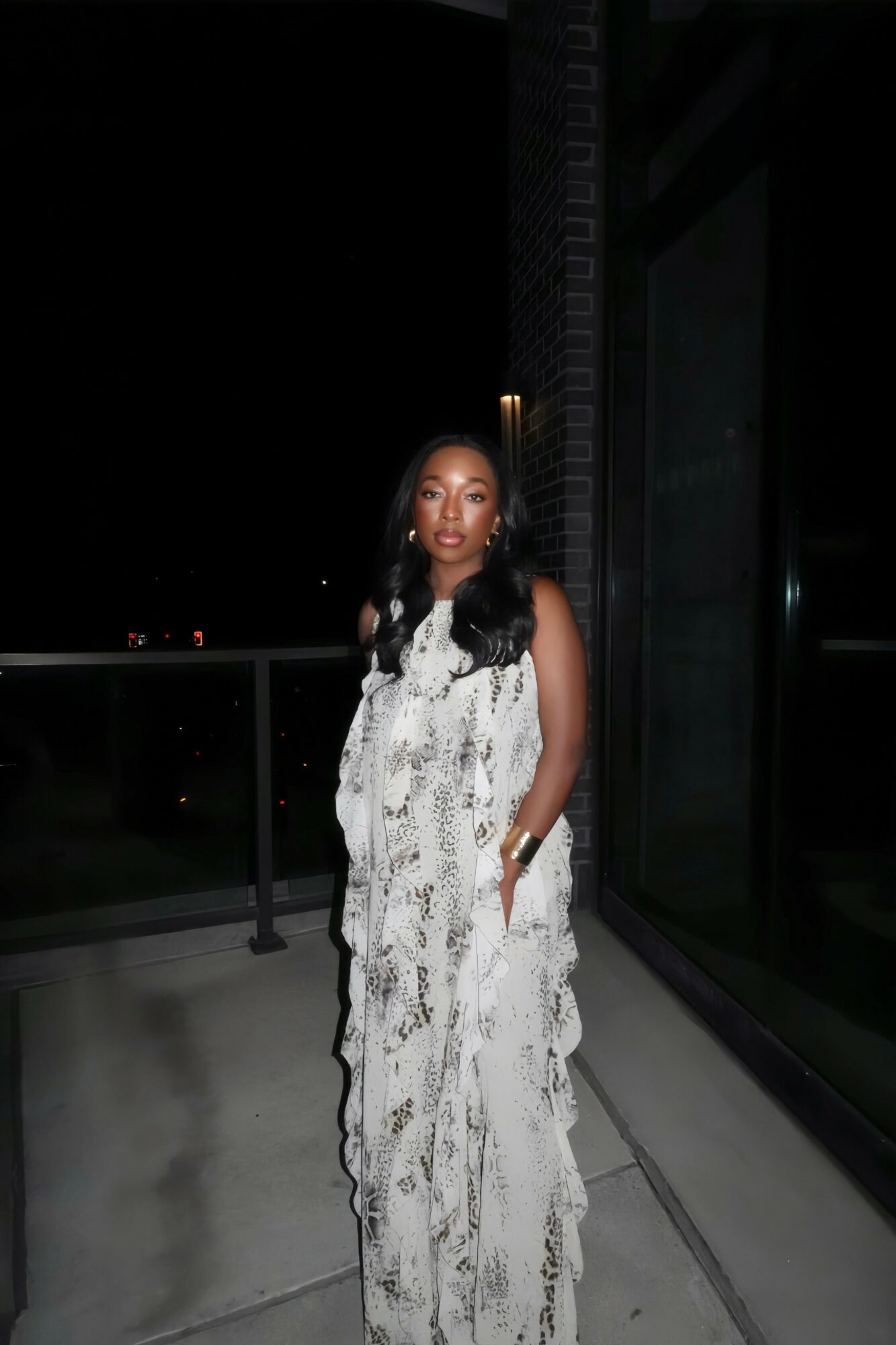 Woman in a long, patterned dress standing on a balcony at night, with glass walls and railing visible.