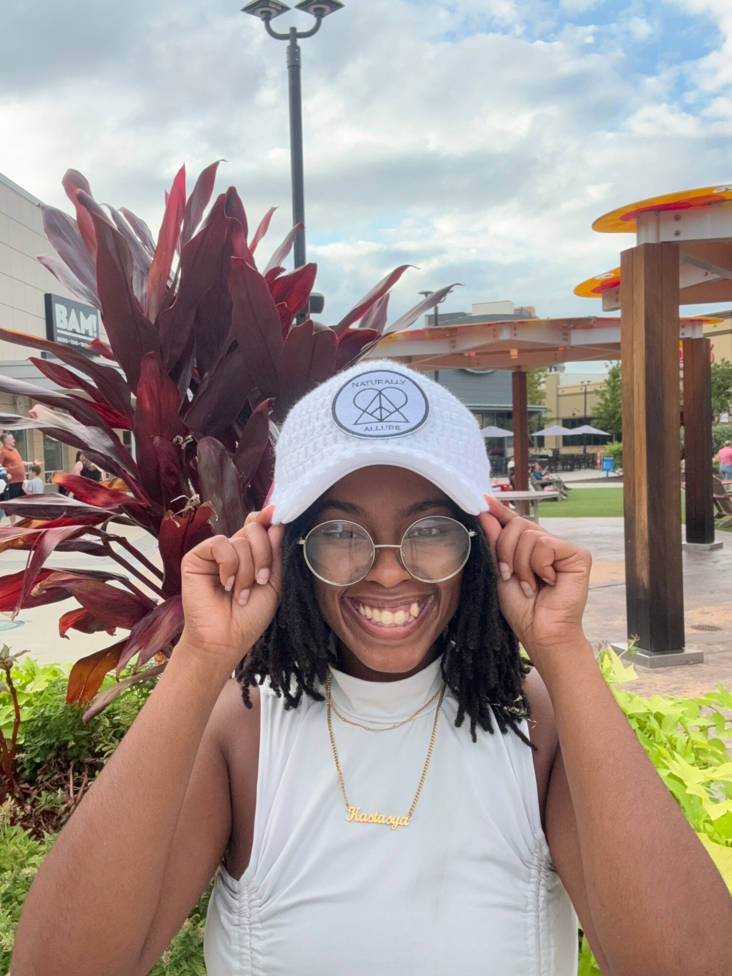 Smiling person with glasses and white cap adjusting glasses outdoors, plants and structures in background.