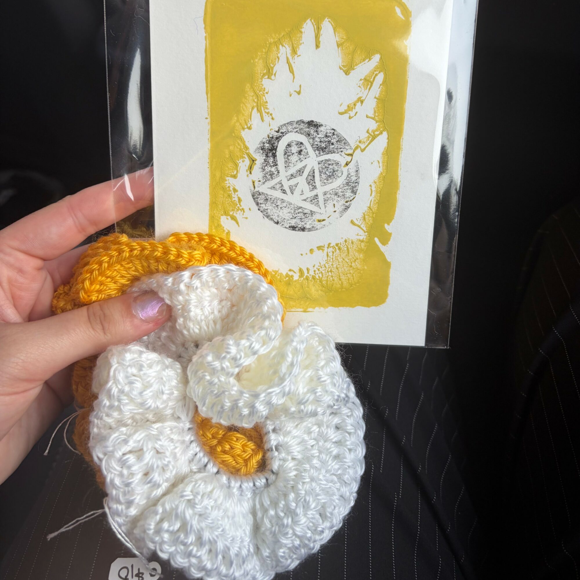 Hand holding a white and yellow crocheted item in front of a yellow and white artwork with a circular design.