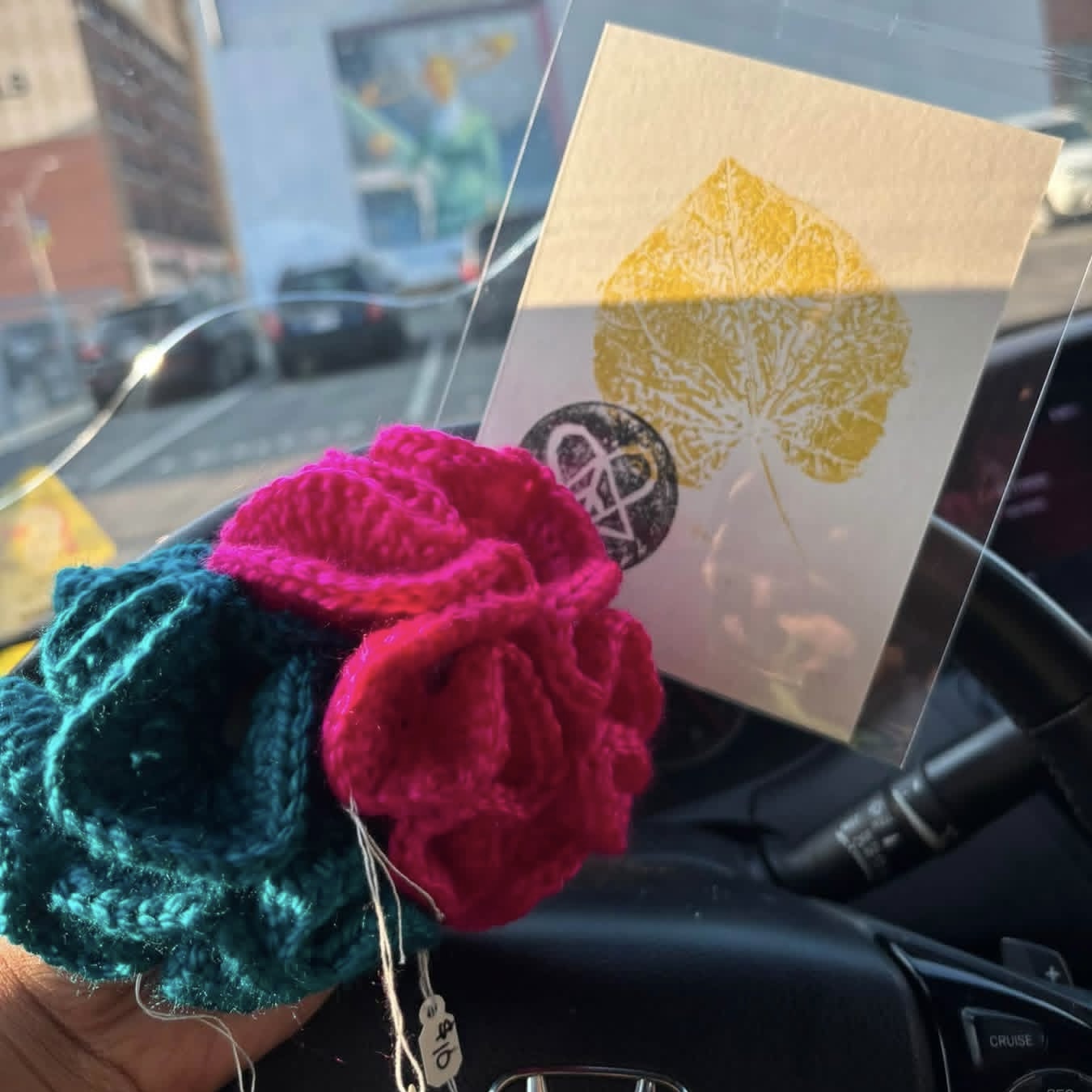 Hand holding colorful knitted item in front of a car dashboard, with a card featuring a yellow leaf and a black and white circle.