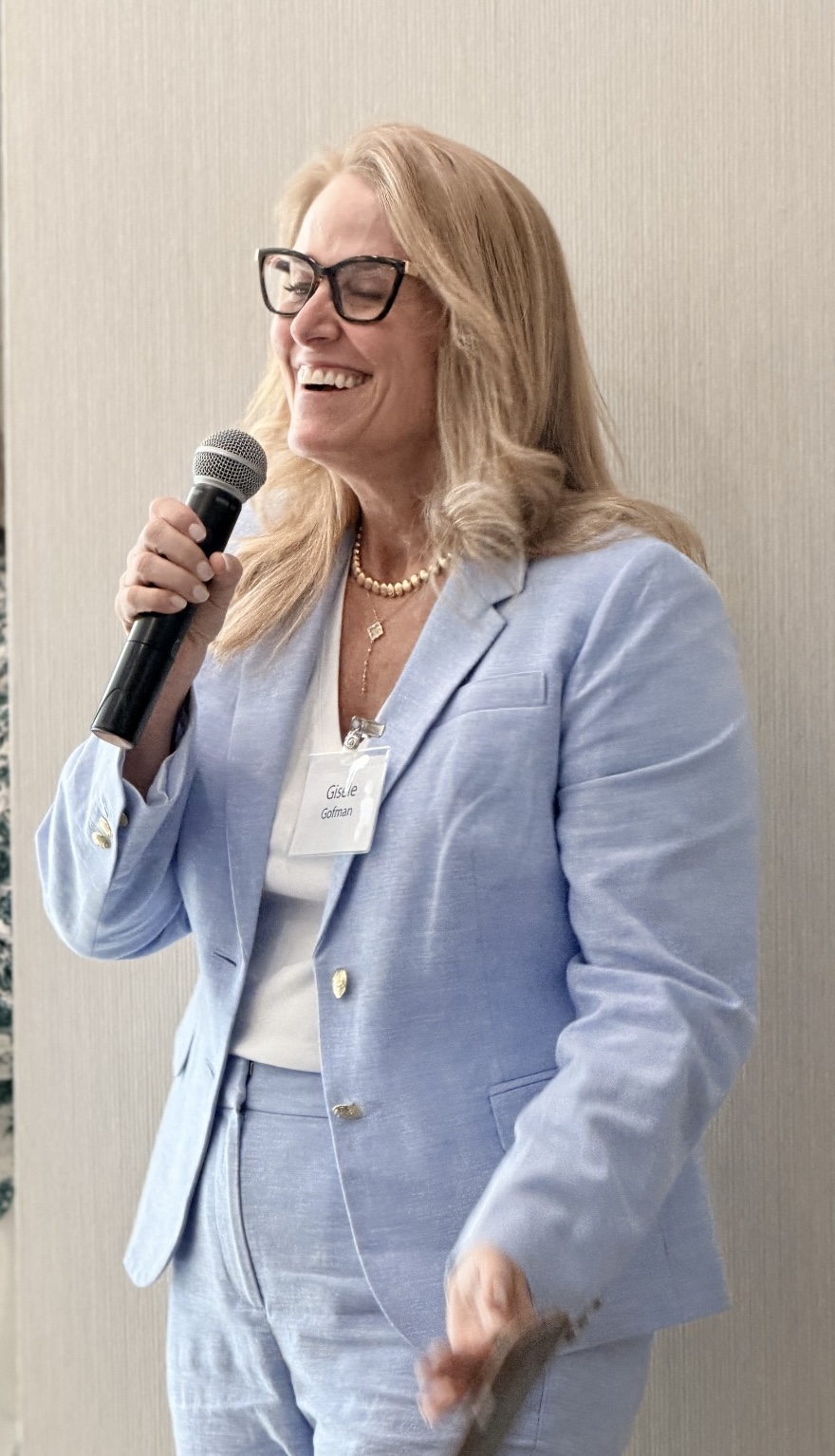 Woman with glasses and blonde hair speaking into a microphone, wearing a light blue blazer and white top.
