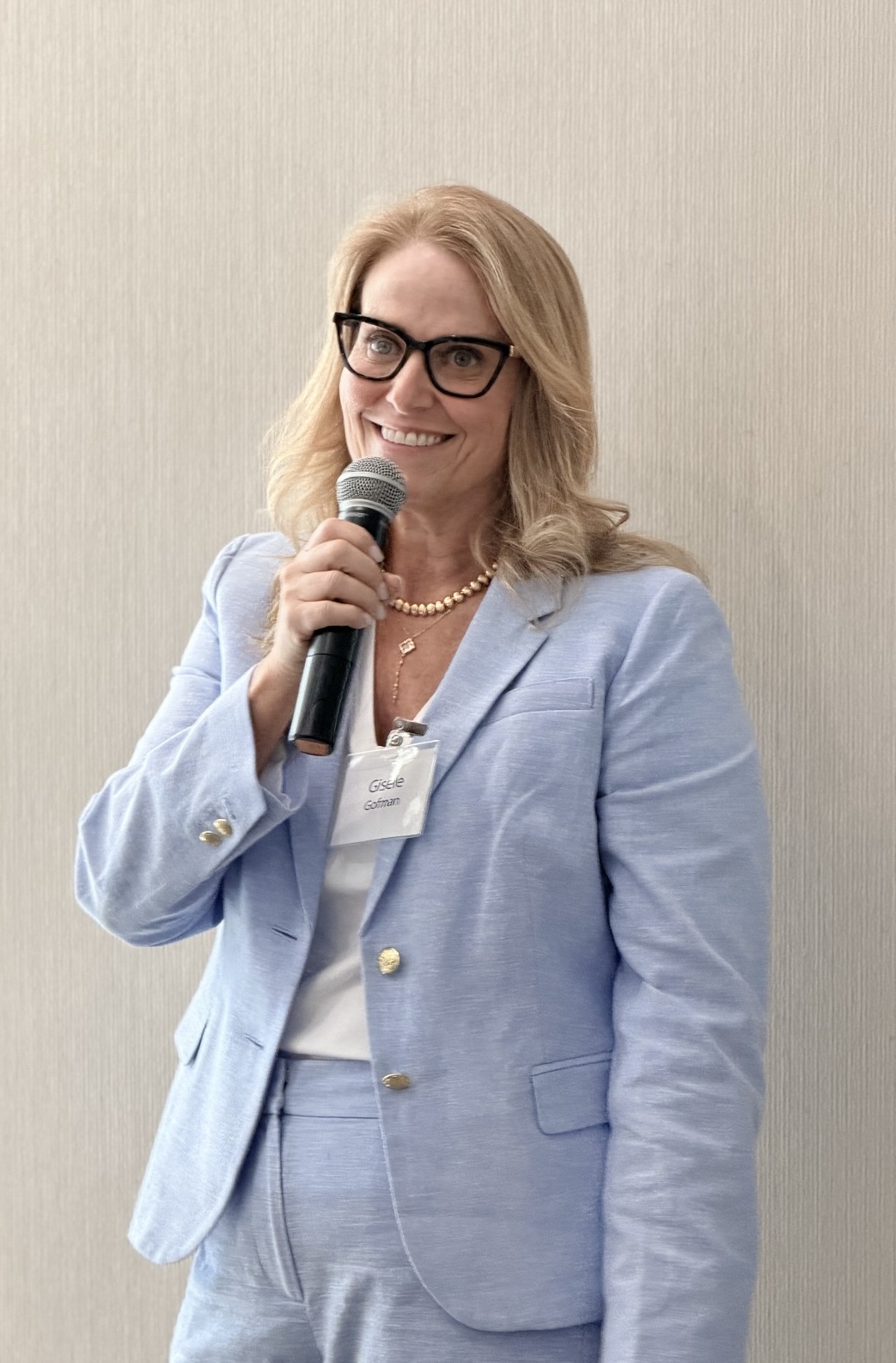 Woman with blonde hair, glasses, and a pearl necklace speaking into a microphone, wearing a light blue suit, smiling.