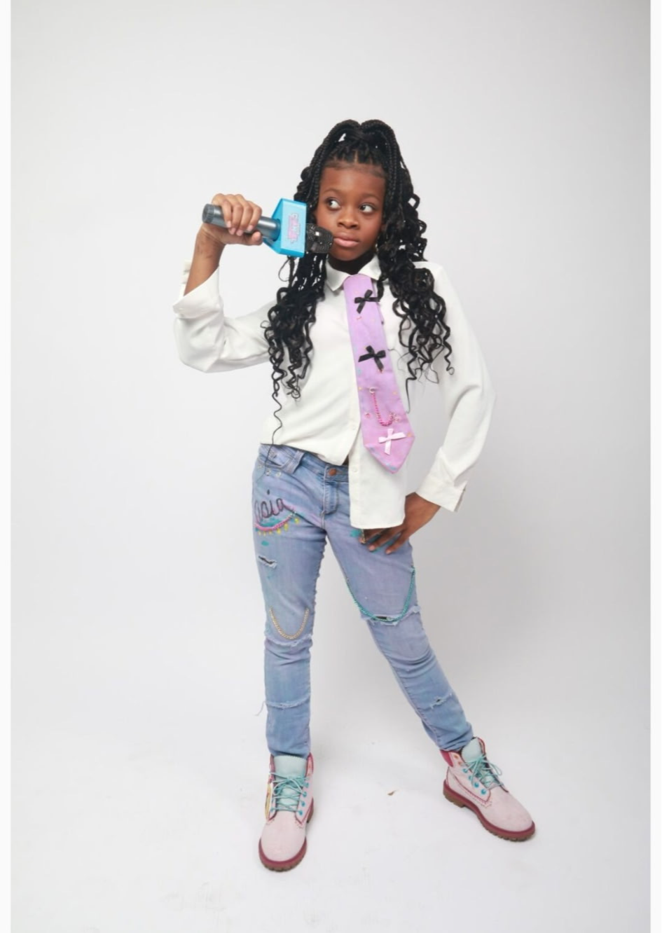 Young girl with long curly hair holding a toy gun, wearing a white jacket, purple backpack, ripped jeans, and sneakers, standing against a plain background.