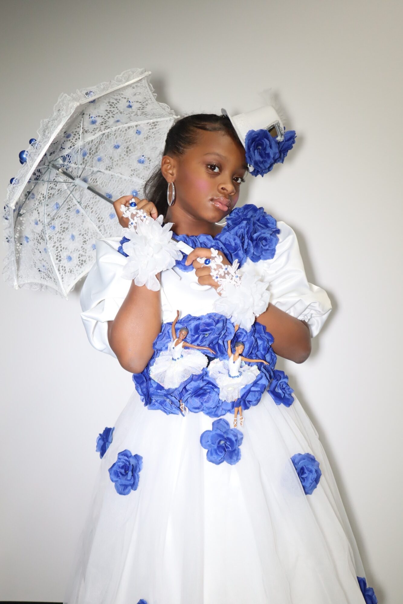 Young girl in a white dress with blue floral accents, holding a transparent umbrella with blue details, against a plain background.