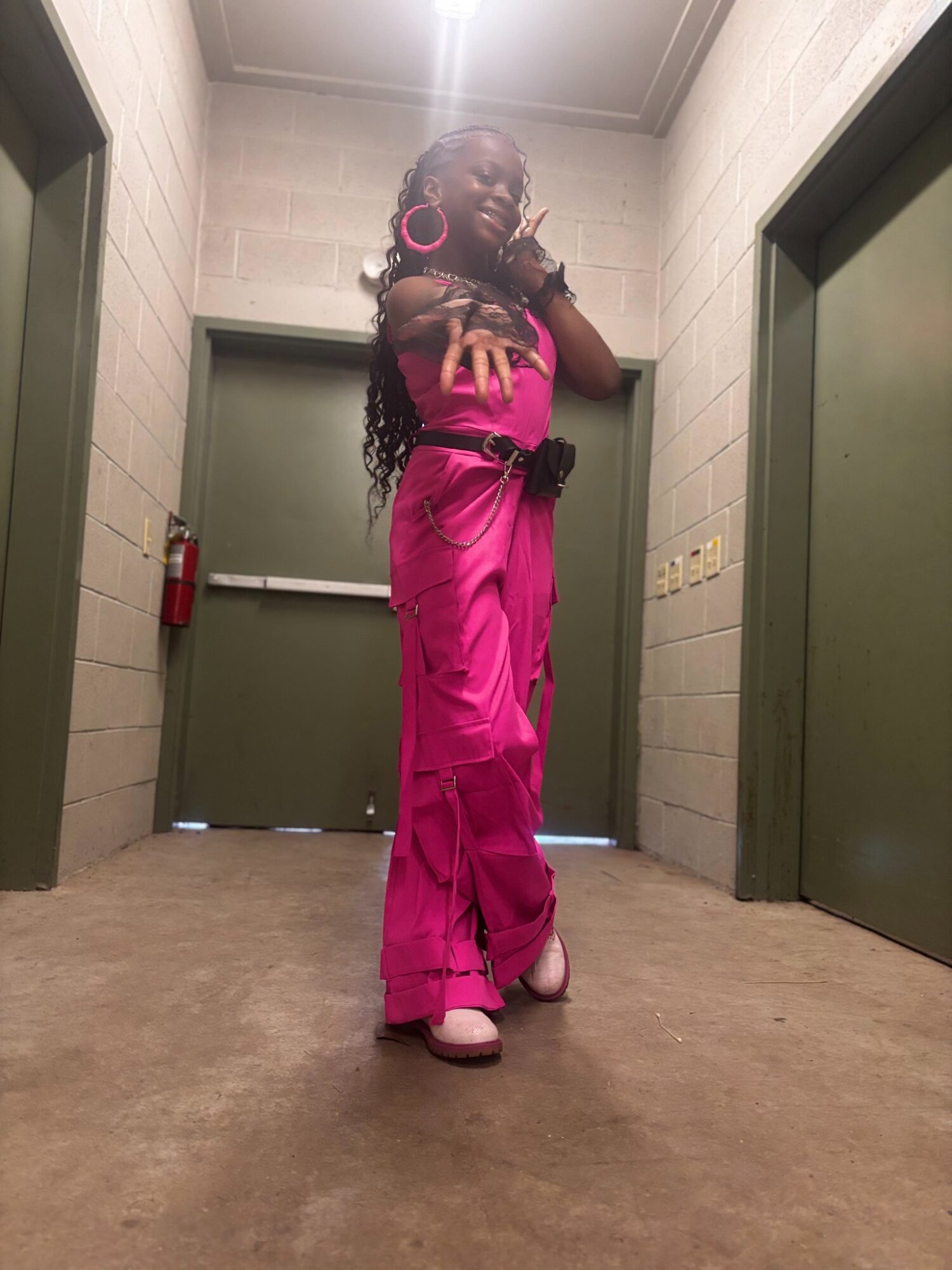 Woman in pink outfit with large earrings standing in a hallway between two green doors, looking to the side.