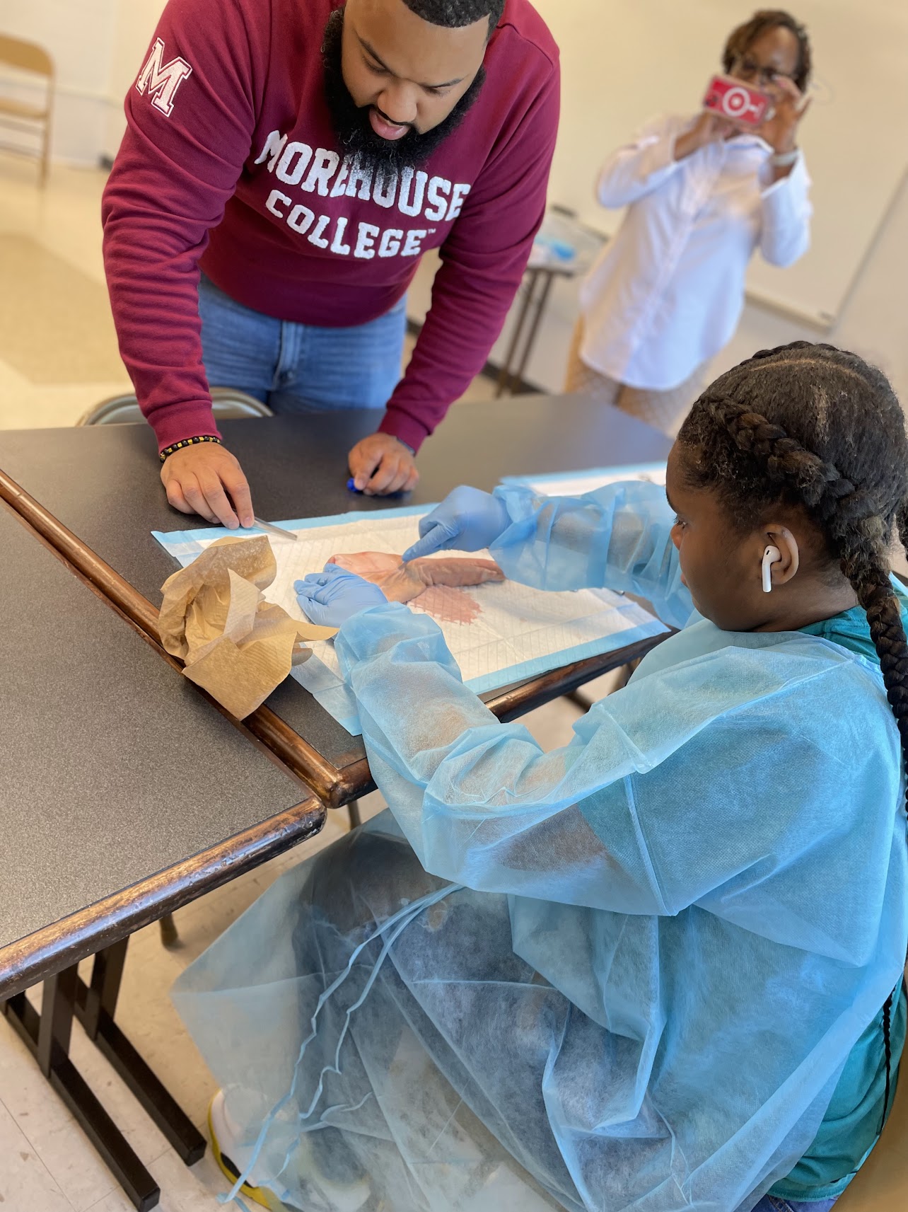 Young girl in a blue protective gown and gloves working with a sample on a table, with a woman and another girl observing in the background.