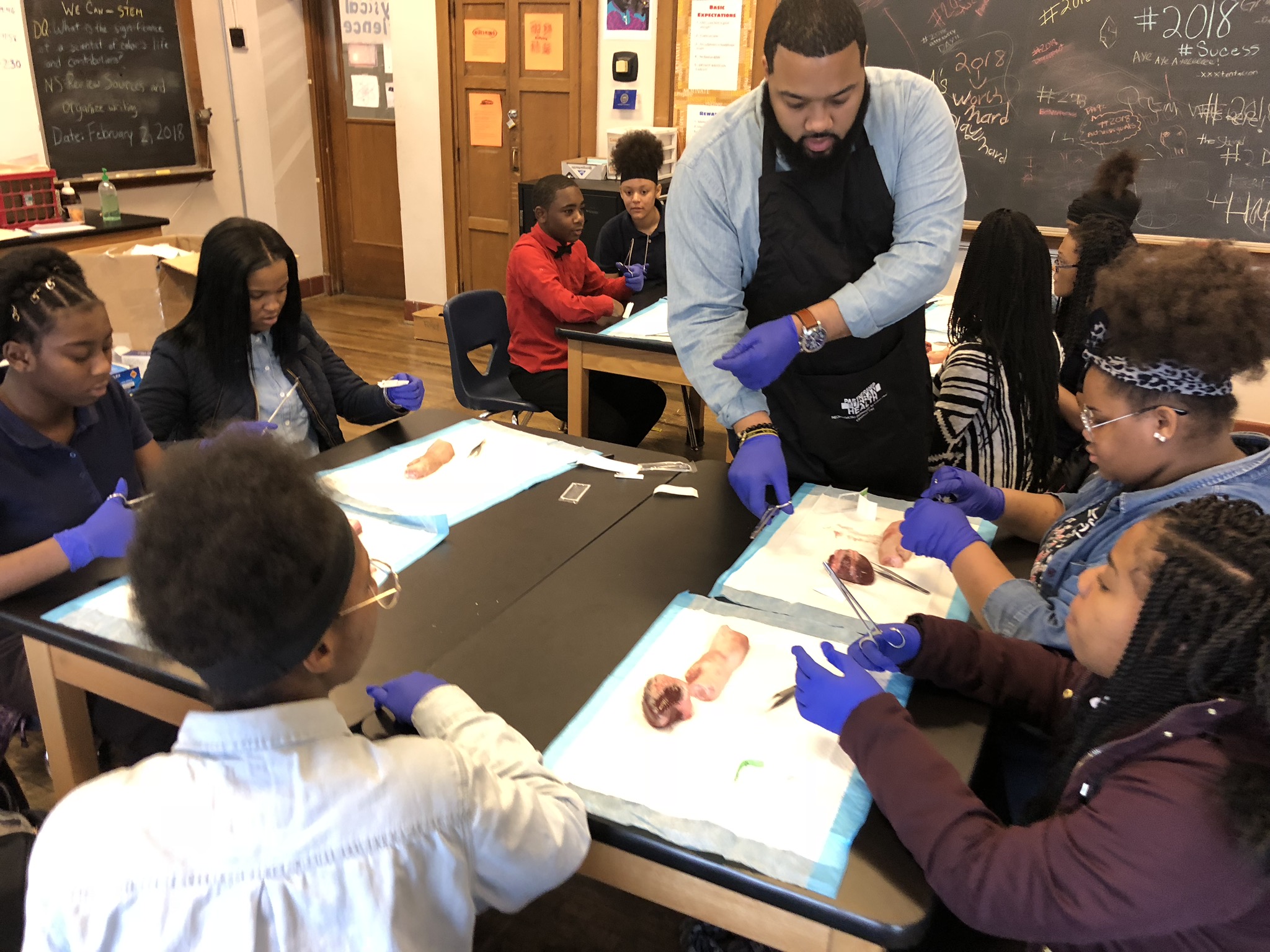 Teacher demonstrating a science experiment to students in a classroom, with students observing and wearing safety gloves.