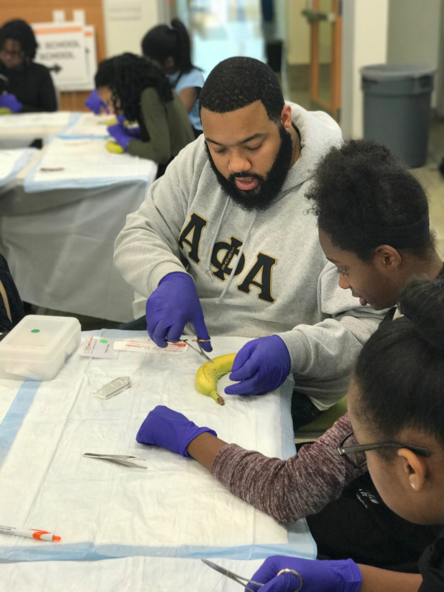 Three people, two women and one man, are working together at a table with yellow objects and papers, wearing purple gloves.