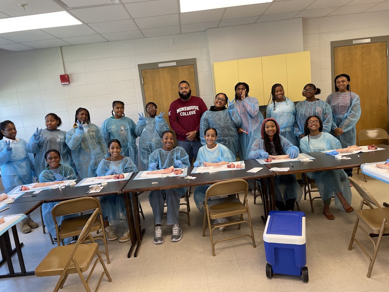 Group of people in blue protective gowns standing and sitting around tables in a room, some smiling, some looking at the camera.