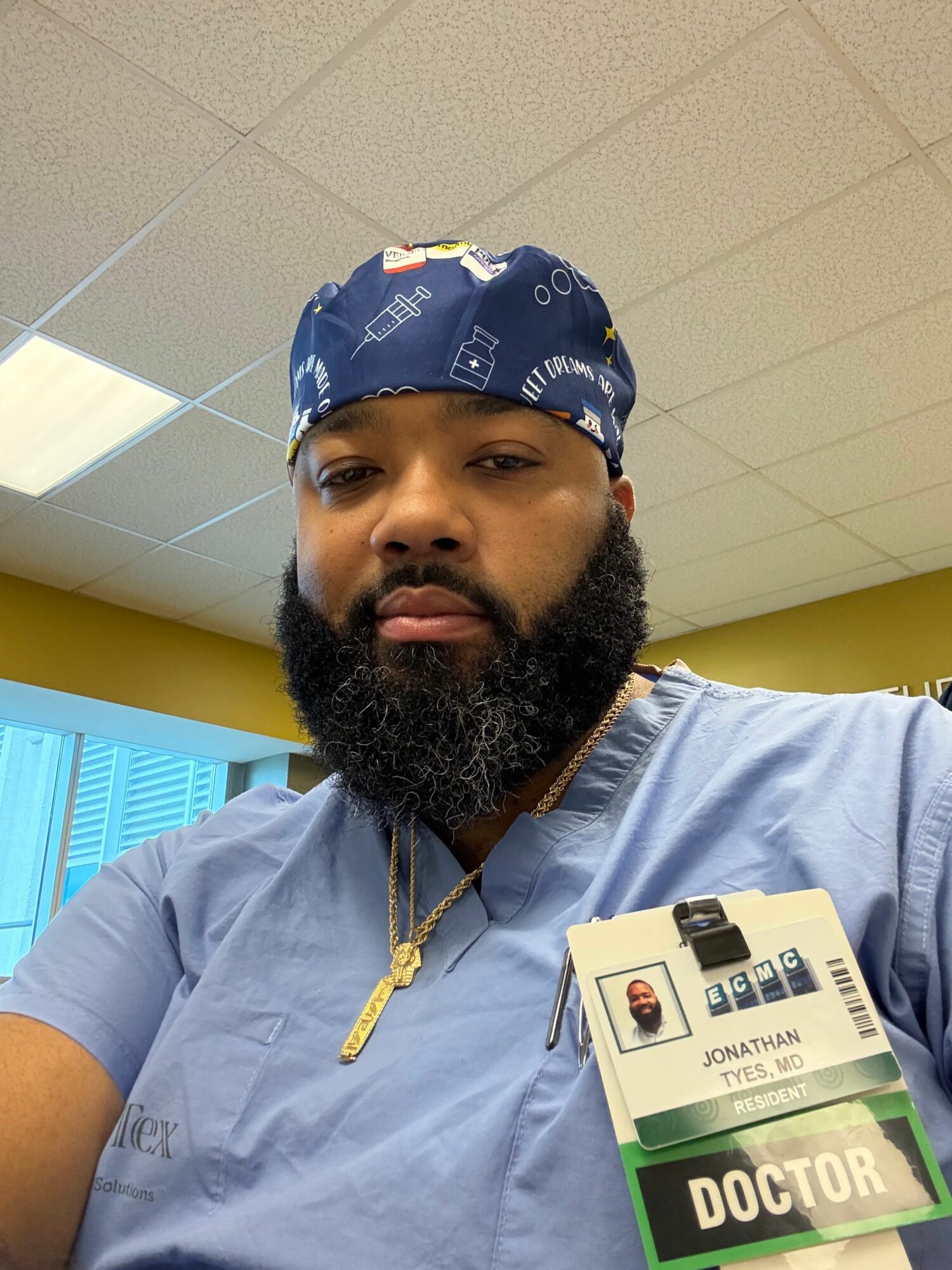 Man with a beard and blue bandana, wearing a blue shirt and ID badge, sitting indoors.