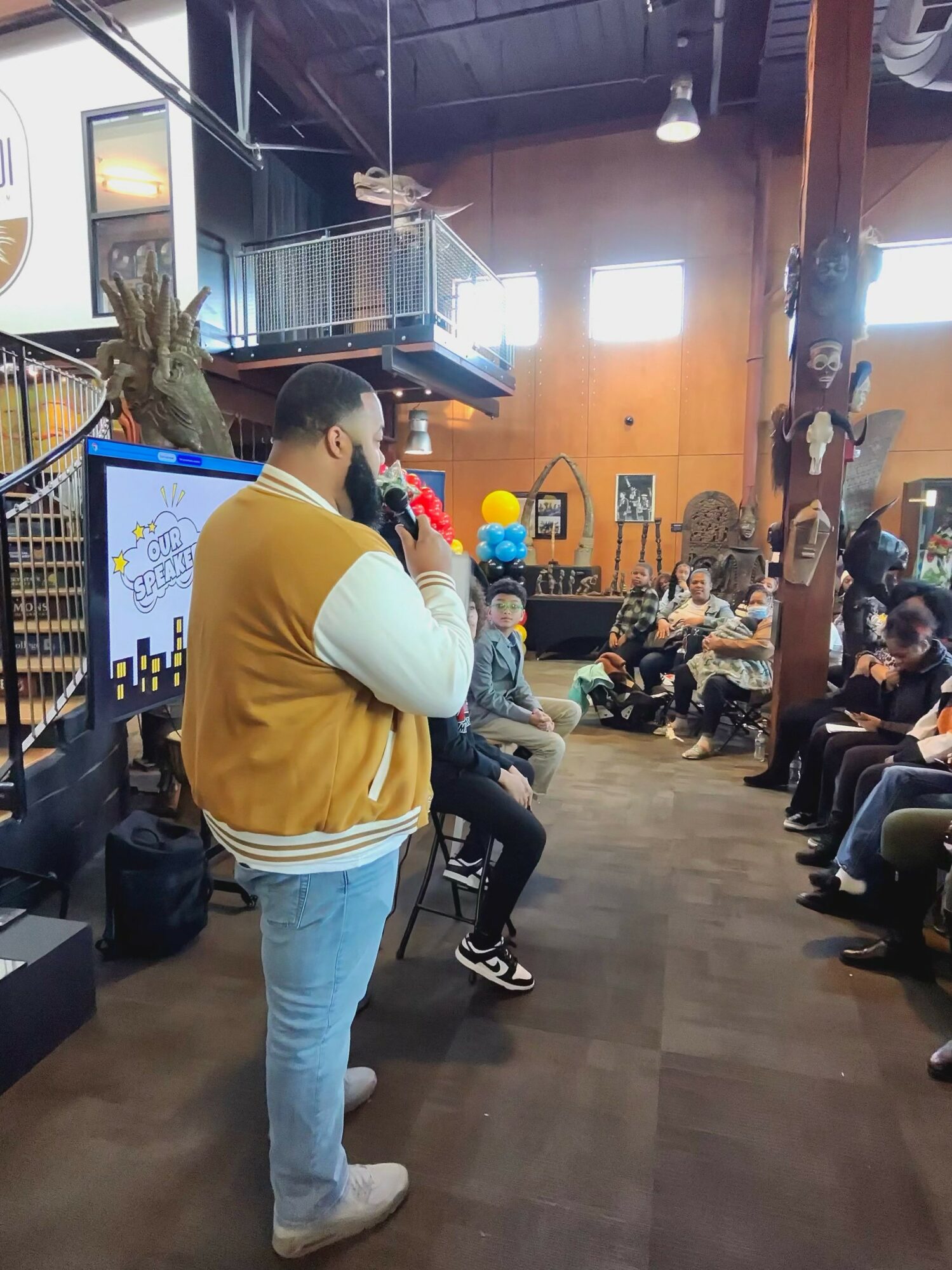 Person in yellow jacket speaking to an audience in a spacious indoor venue with colorful decorations and people seated.
