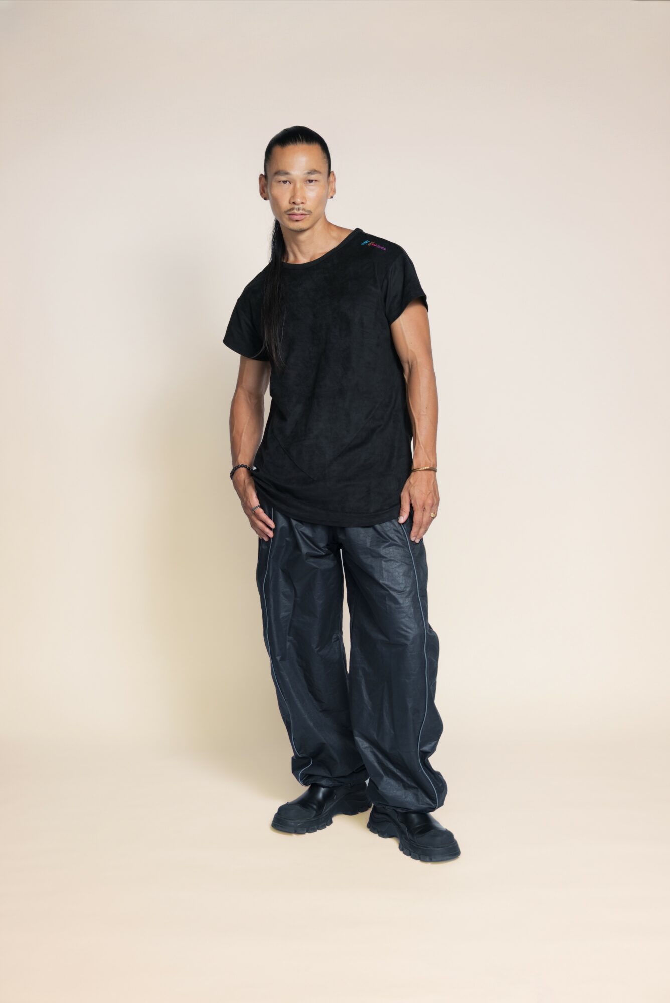 Young man standing against a plain background, wearing a black t-shirt and baggy jeans, looking at the camera.