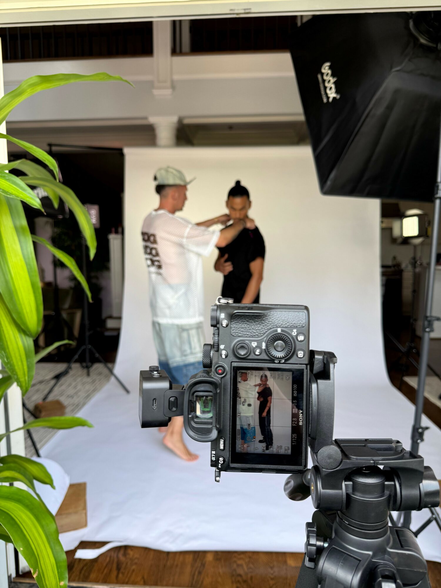 Two people in a studio with a camera on a tripod, one person standing and the other sitting, plants in foreground.