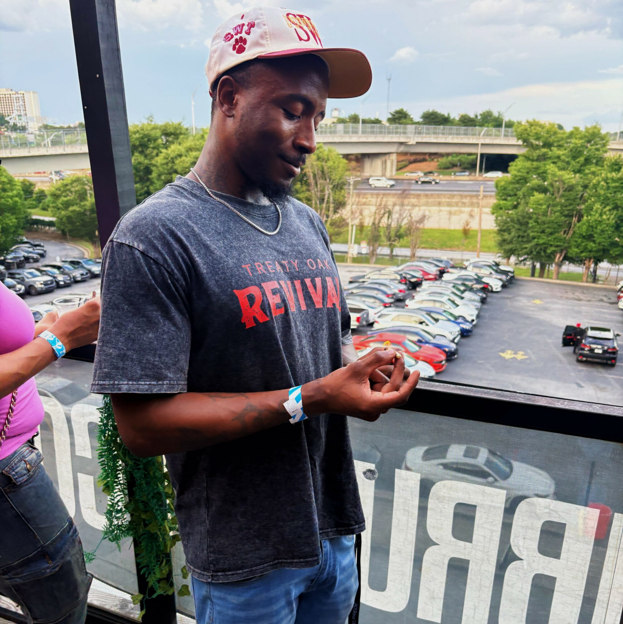 Young man wearing a cap and T-shirt, standing outdoors near a railing, with a parking lot and trees in the background.