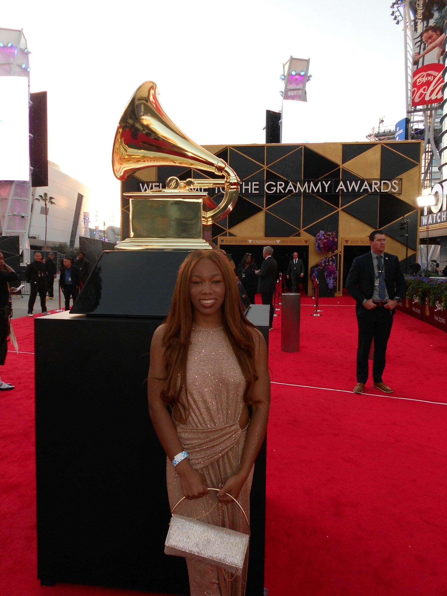 Young woman with long hair in a beige dress holding a purse on a red carpet at the Grammy Awards, large golden gramophone statue behind her.