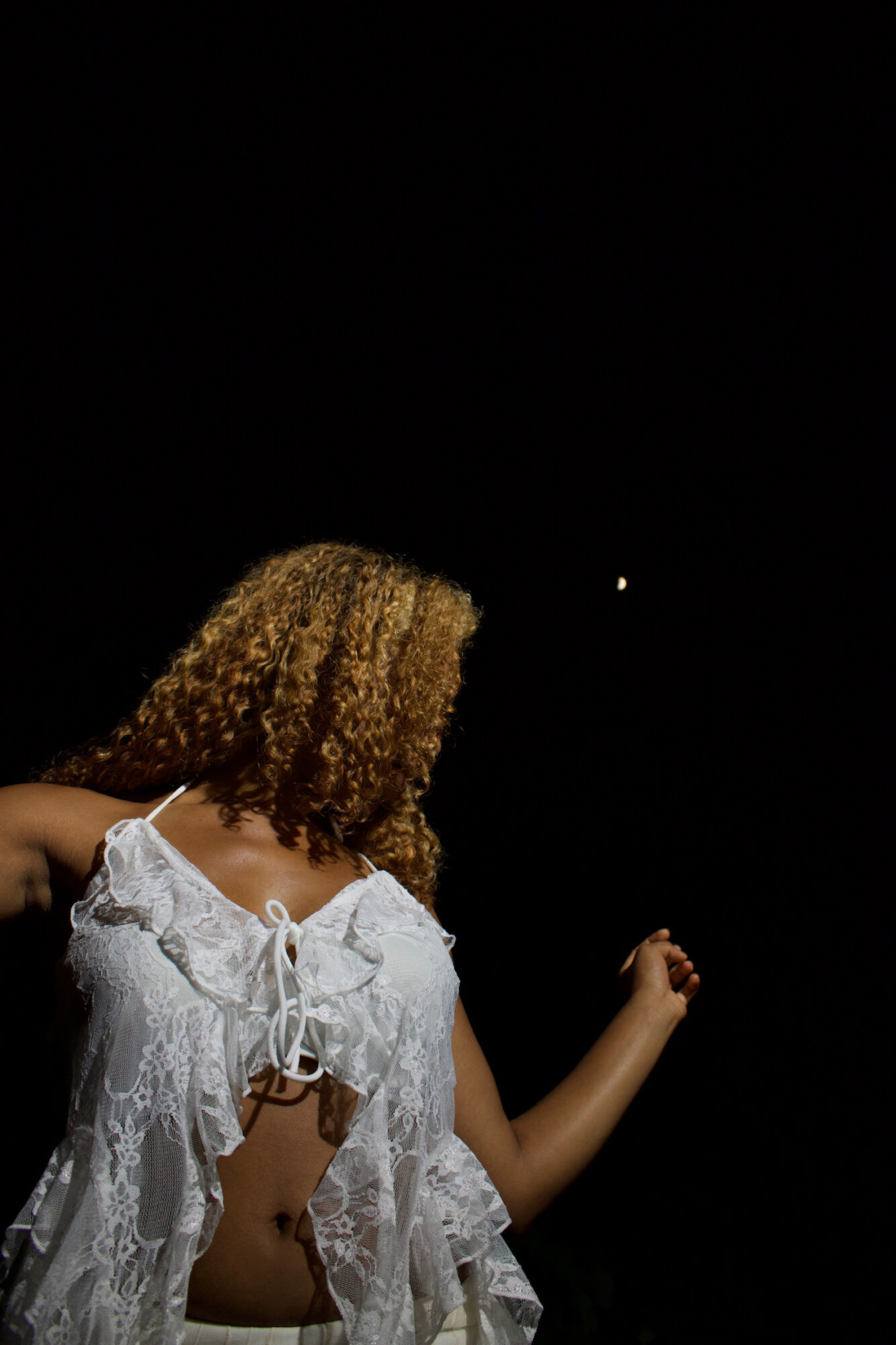 Person with curly hair wearing a white lace top, facing away in a dark background with a small light in the distance.
