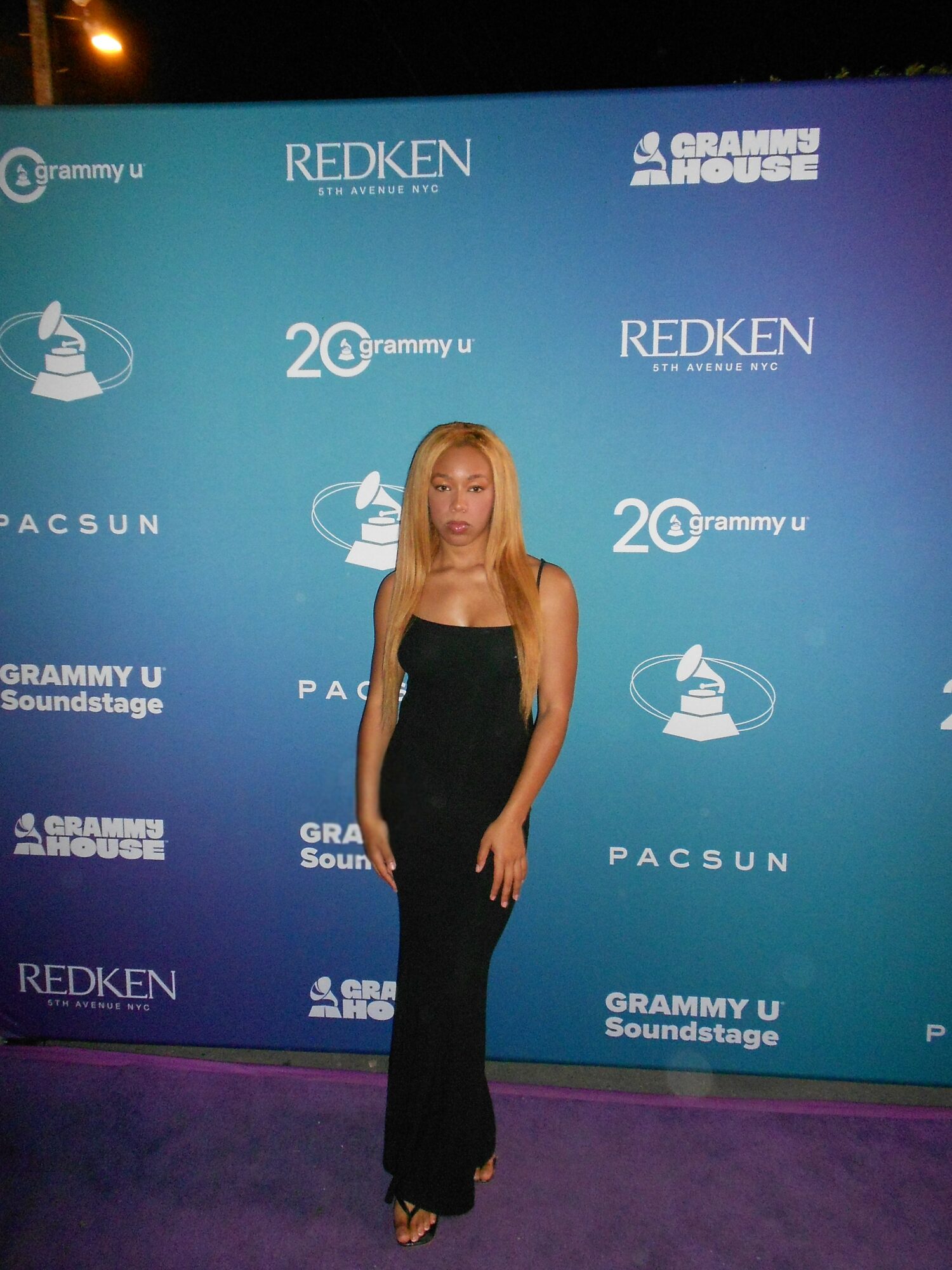 Woman in a black dress standing in front of a blue backdrop with logos and text, including Grammy U and Redken.