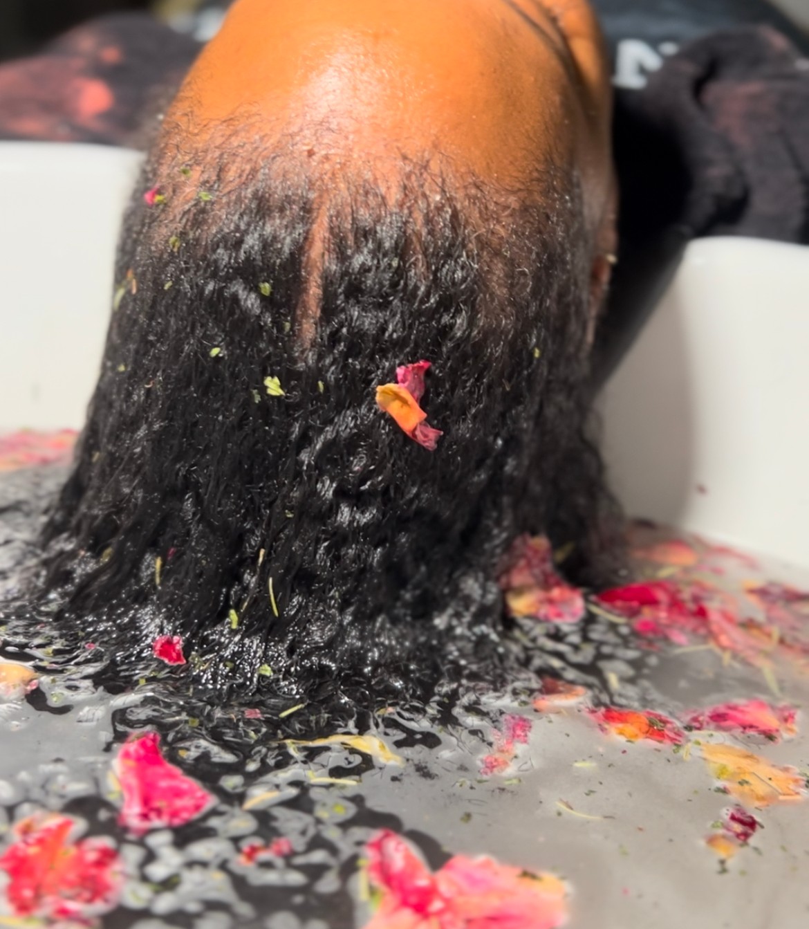 Person's head with wet, curly hair covered in flower petals and debris, leaning forward over a surface with scattered petals.