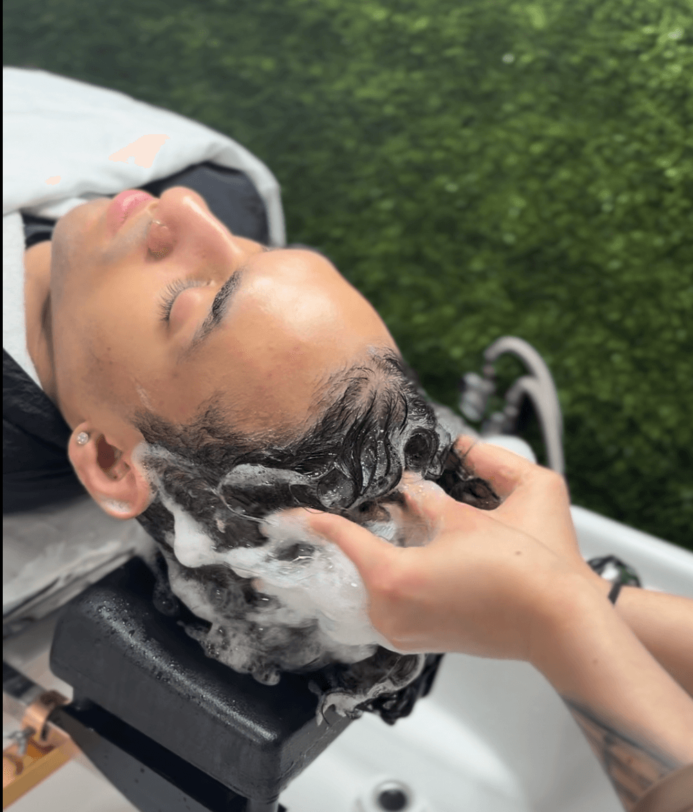 Person receiving a head wash with soap lather, lying on a chair outdoors with greenery in the background.