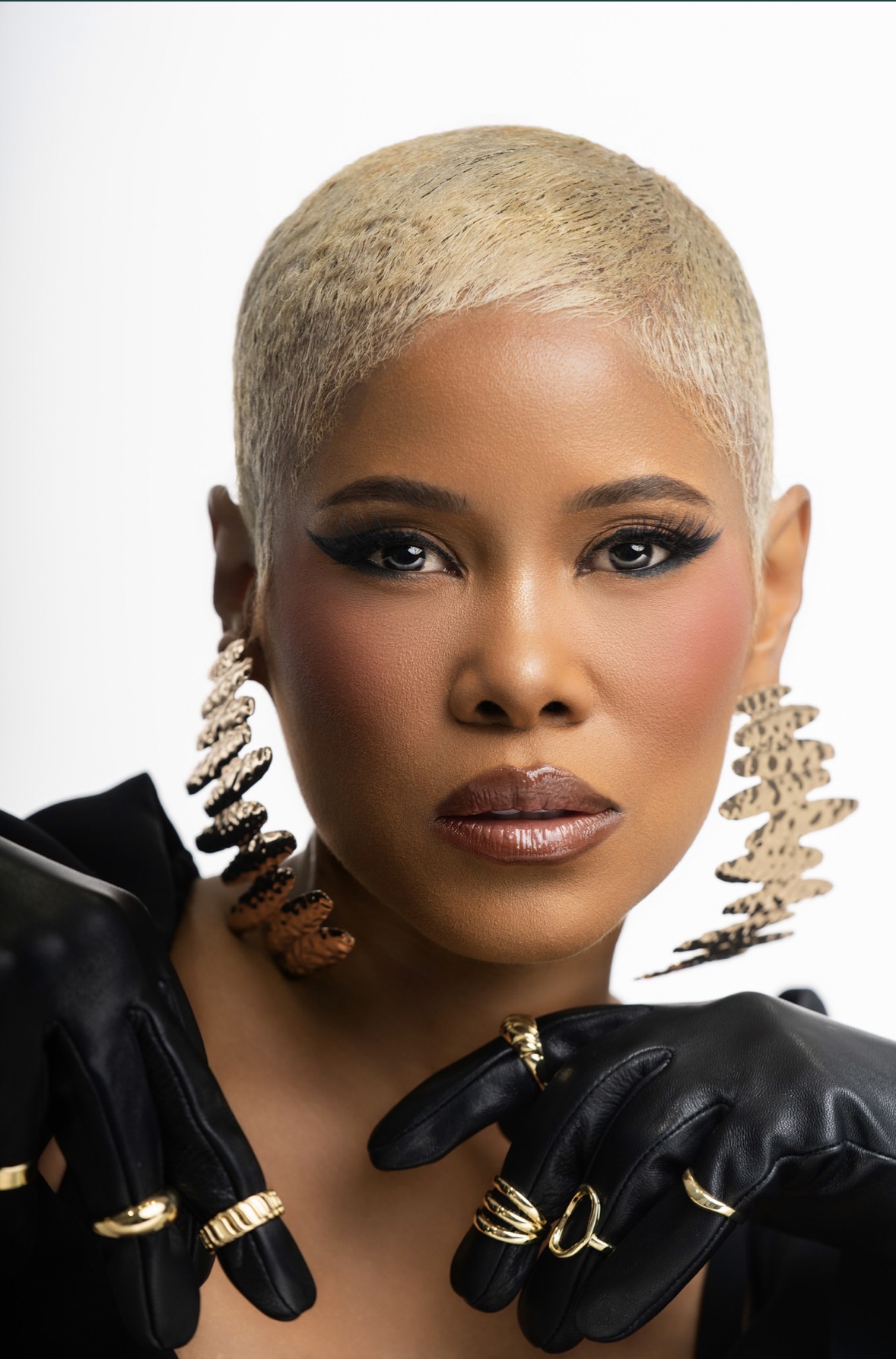 Woman with short blonde hair, bold makeup, large earrings, and black gloves, looking directly at the camera.