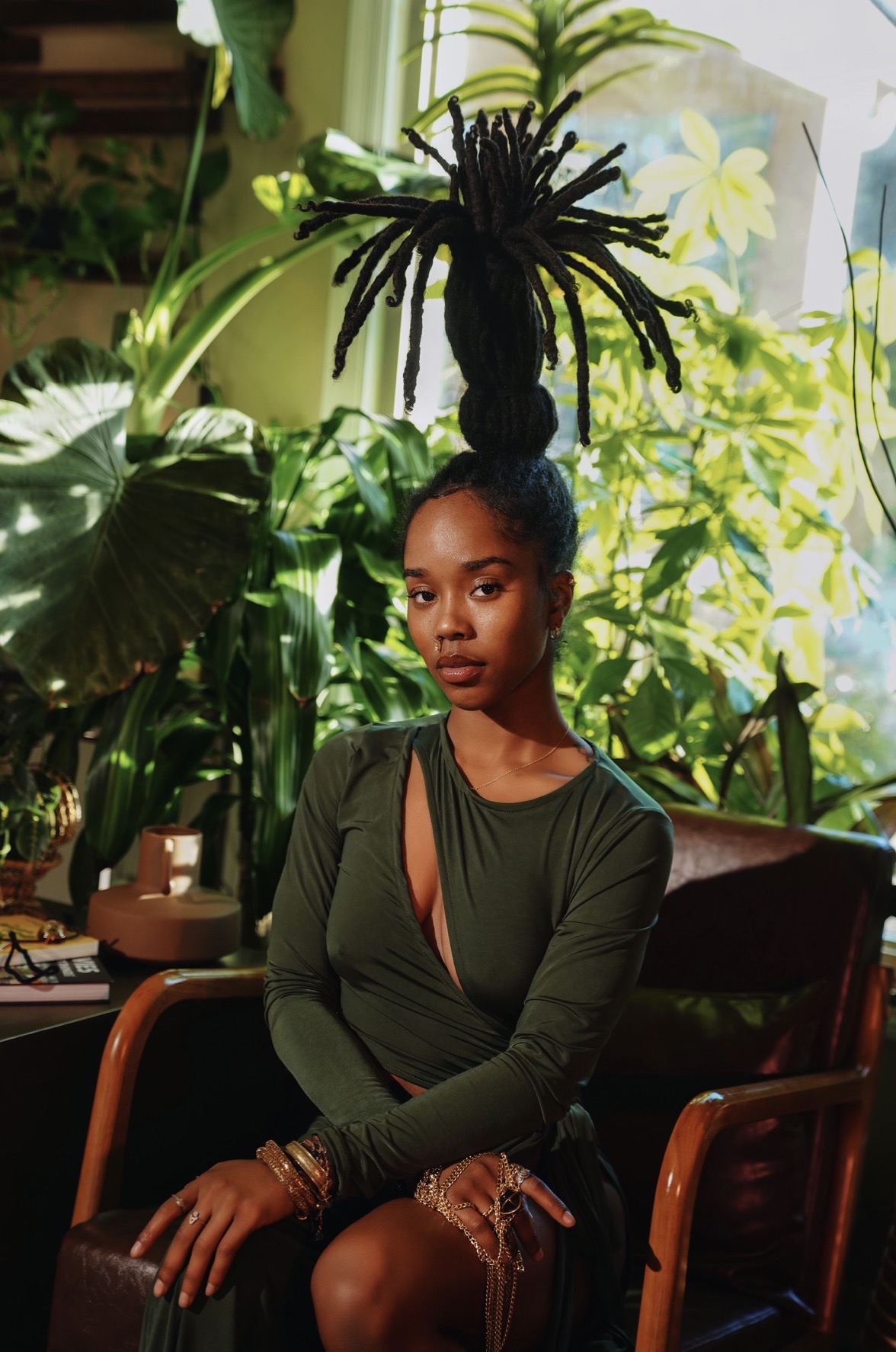 Young woman with dreadlocks styled in a high bun, sitting on a wooden chair surrounded by lush green plants.