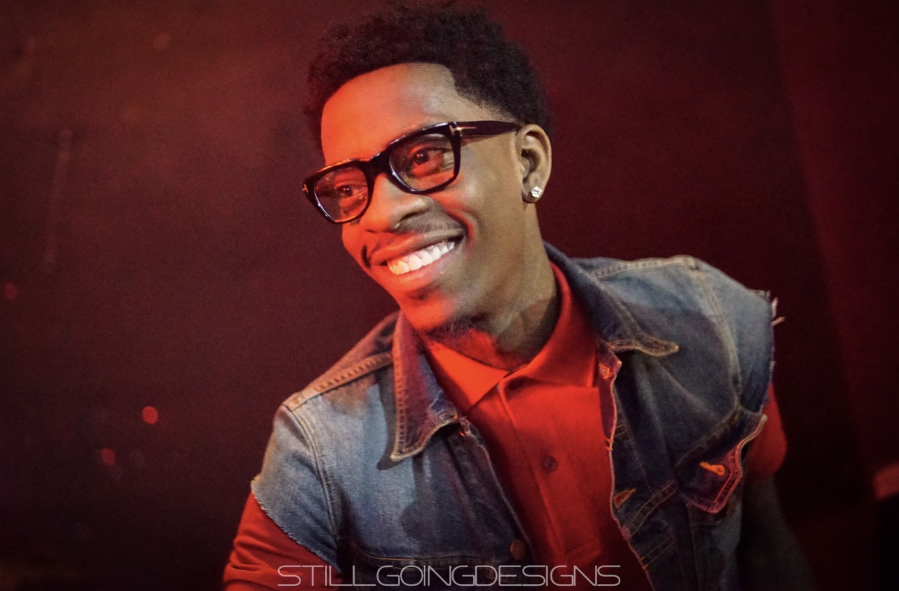 Young man smiling, wearing glasses, red shirt, and denim jacket, against a dark background.