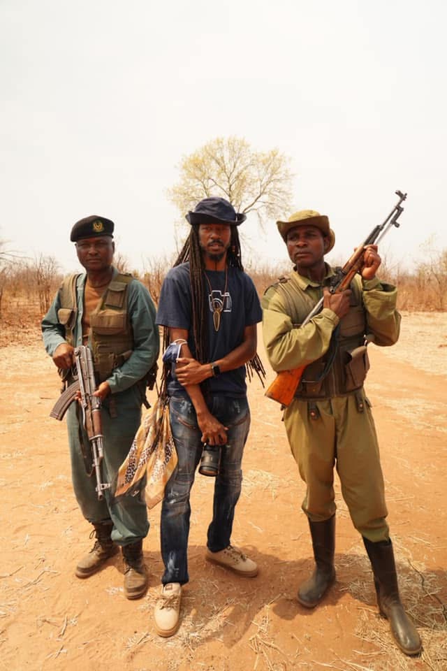 Three men standing outdoors, two holding rifles, one with long dreadlocks, in a dry landscape with a tree in the background.