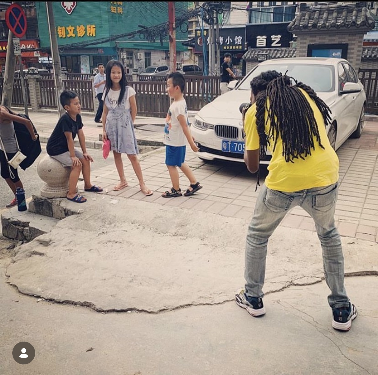 Group of children and a woman on sidewalk near a white car, with a man taking photo, urban background.