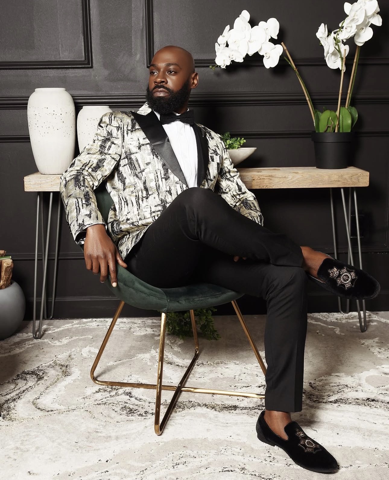 Man in patterned blazer and black pants sitting on a green chair in a room with dark walls and white orchids.