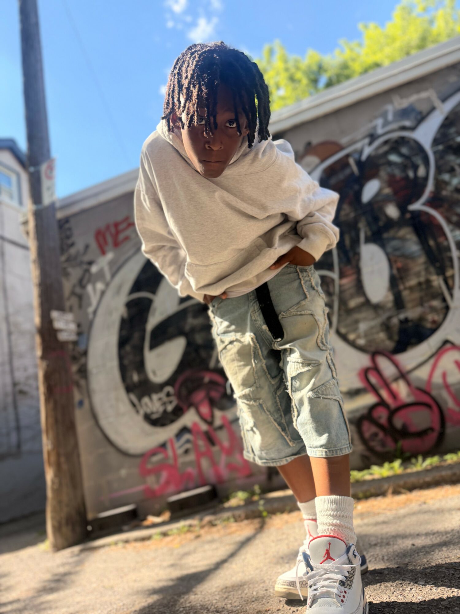 Child with dreadlocks wearing a white hoodie, ripped jeans, and sneakers standing outdoors near graffiti wall.