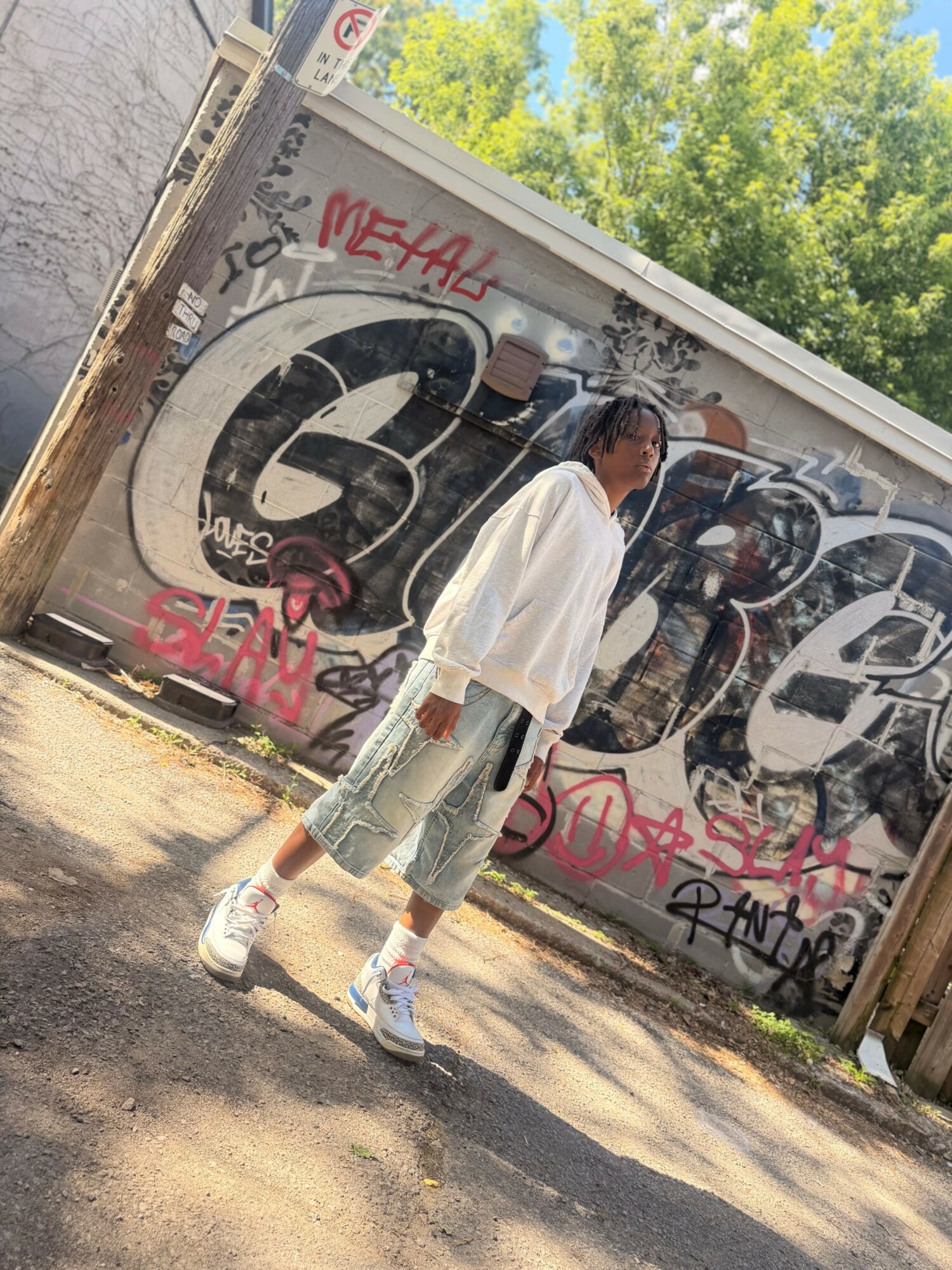 Person with white shirt and jeans standing in front of graffiti-covered wall, trees in background.
