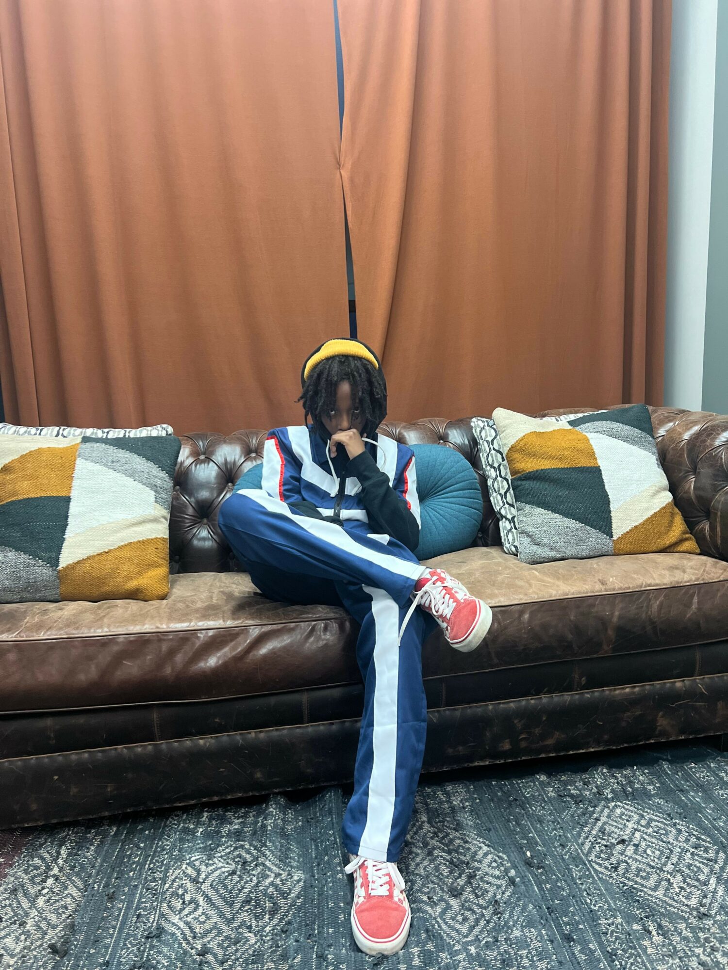 Child sitting on a sofa with colorful pillows, wearing a striped jacket and sneakers, in a room with curtains and a patterned rug.