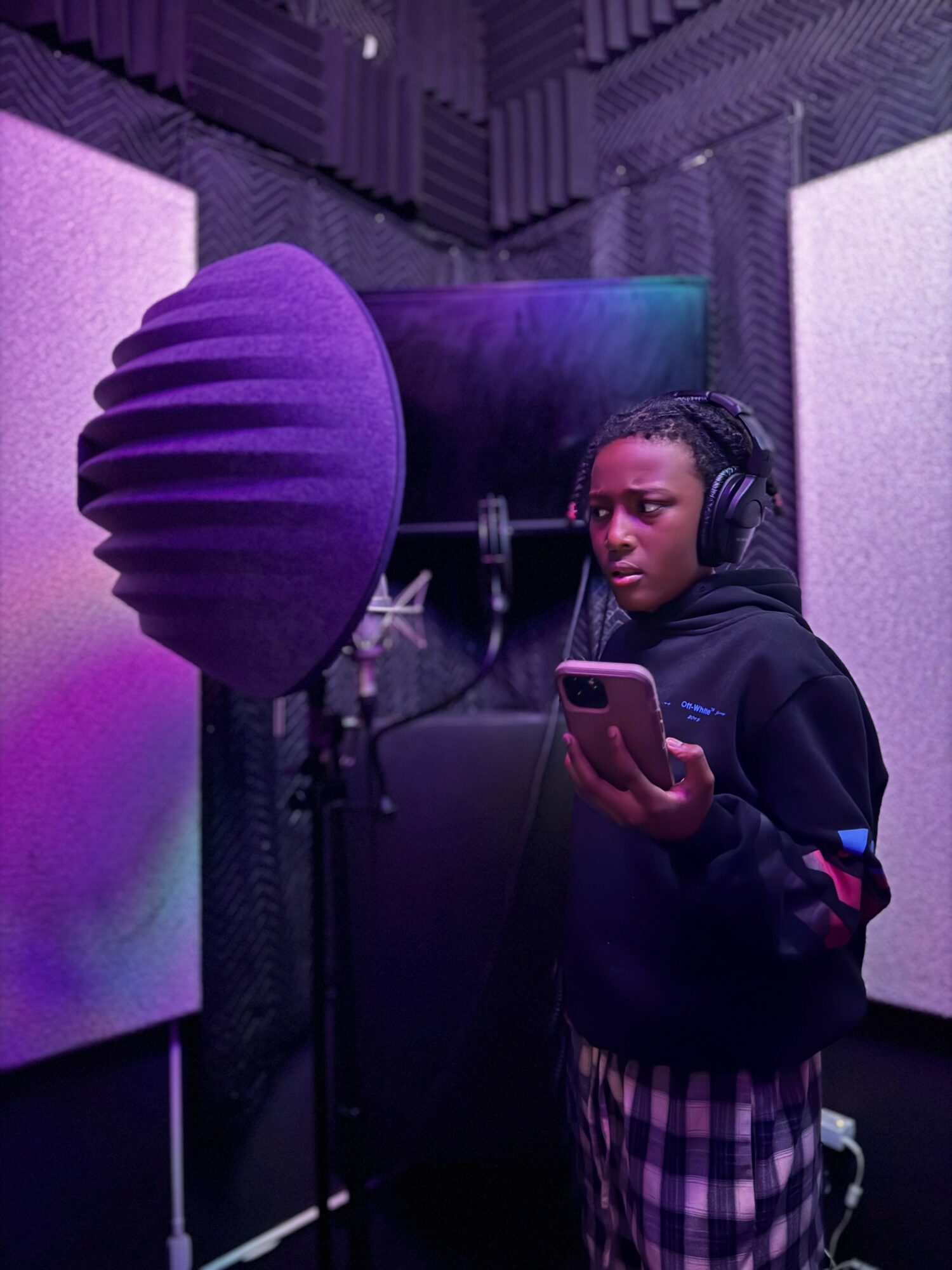Young person in a recording studio with headphones, holding a phone, purple soundproofing panels, and a microphone with a pop filter.