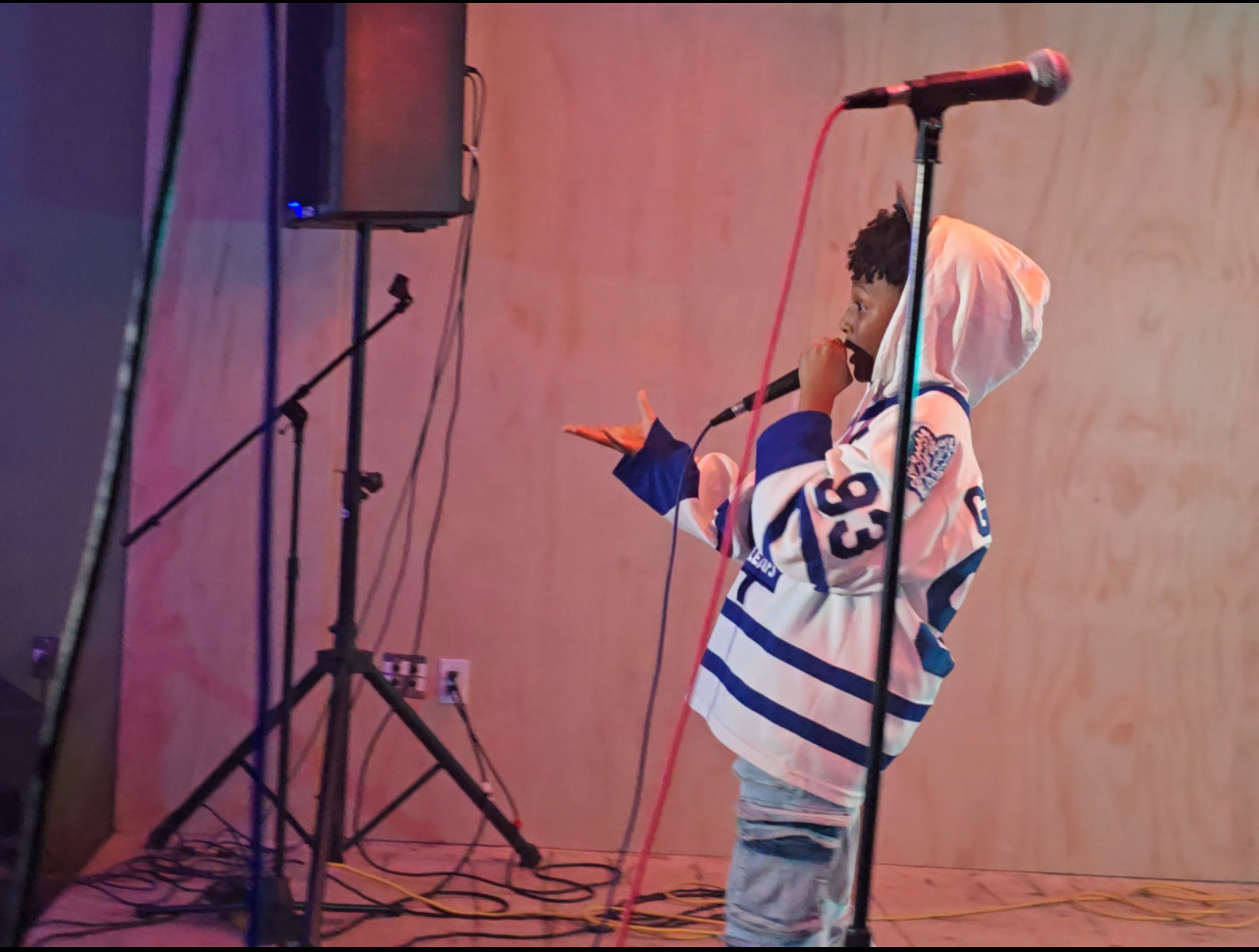 Child with curly hair in a hoodie and striped shirt holding a microphone, standing near a microphone stand on stage.