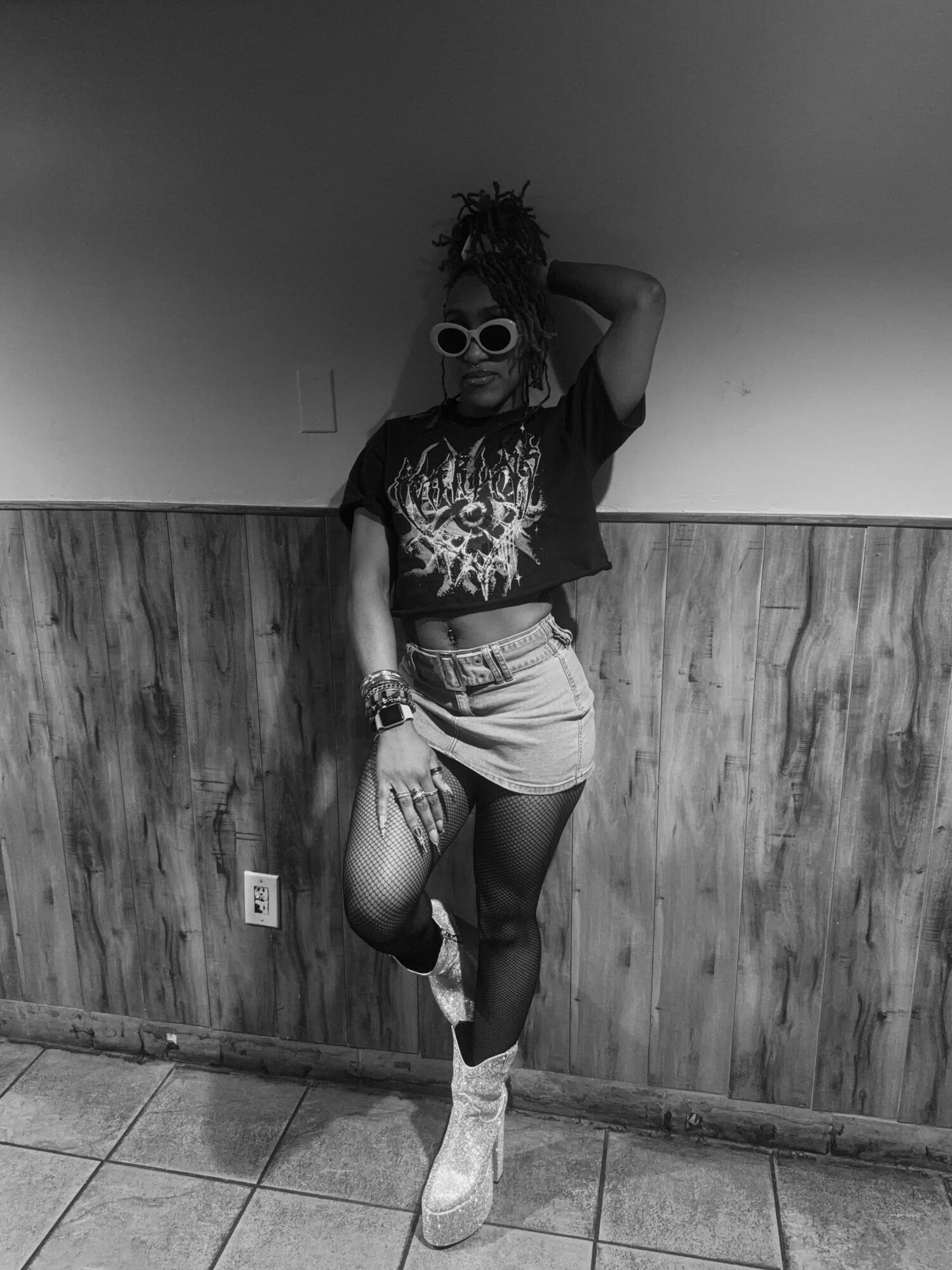 Person with dreadlocks wearing sunglasses, a graphic t-shirt, a skirt, and sneakers, posing with one leg raised and hand behind head.
