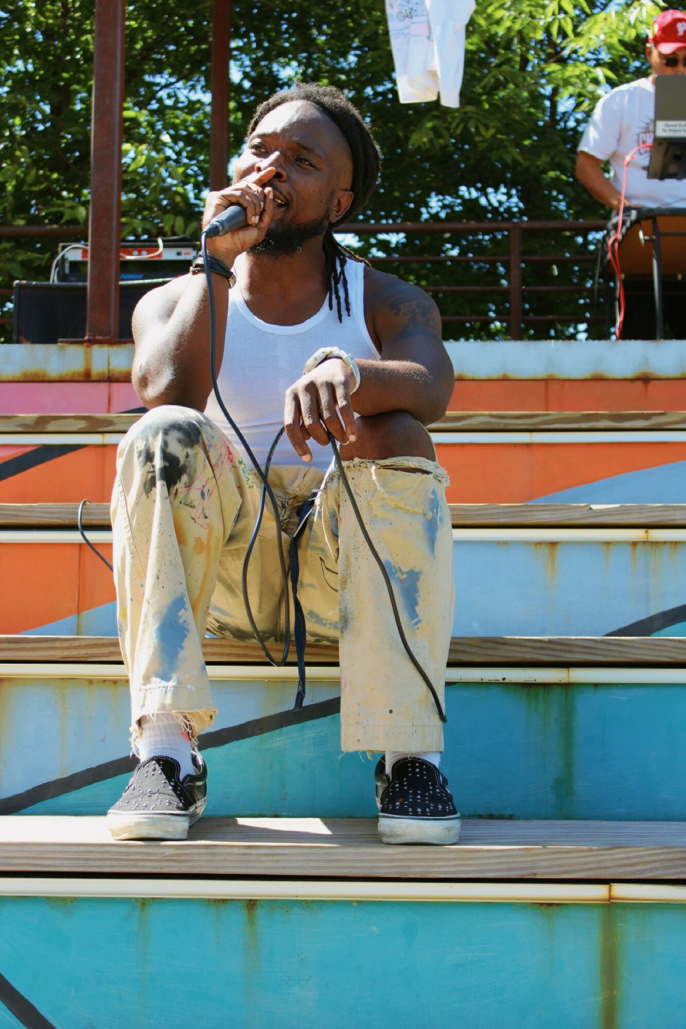Young man with dreadlocks singing into a microphone outdoors, sitting on steps with trees and people in background.