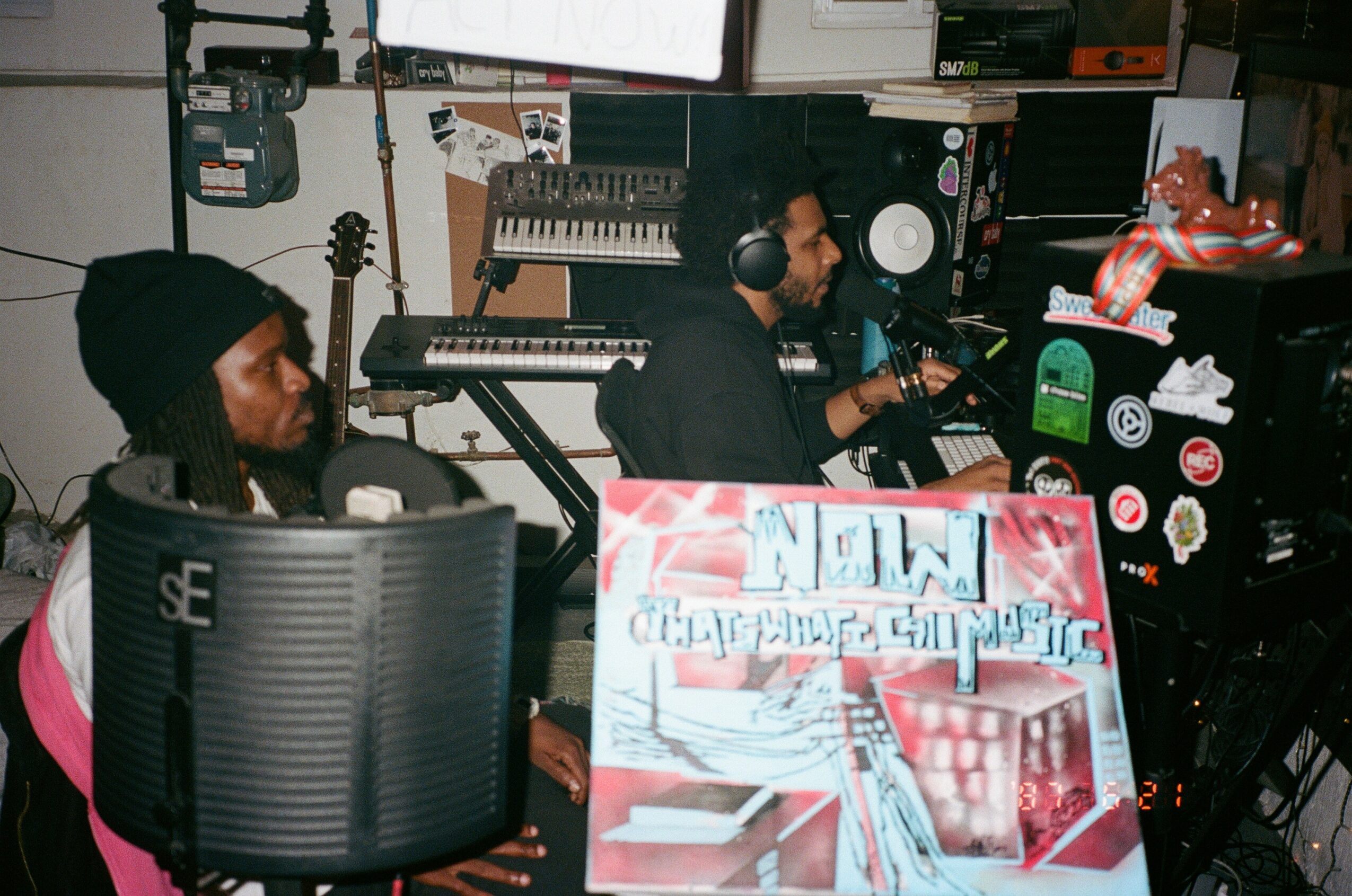 Two people in a music studio with equipment, one speaking into a microphone, a keyboard, and a colorful sign in the foreground.