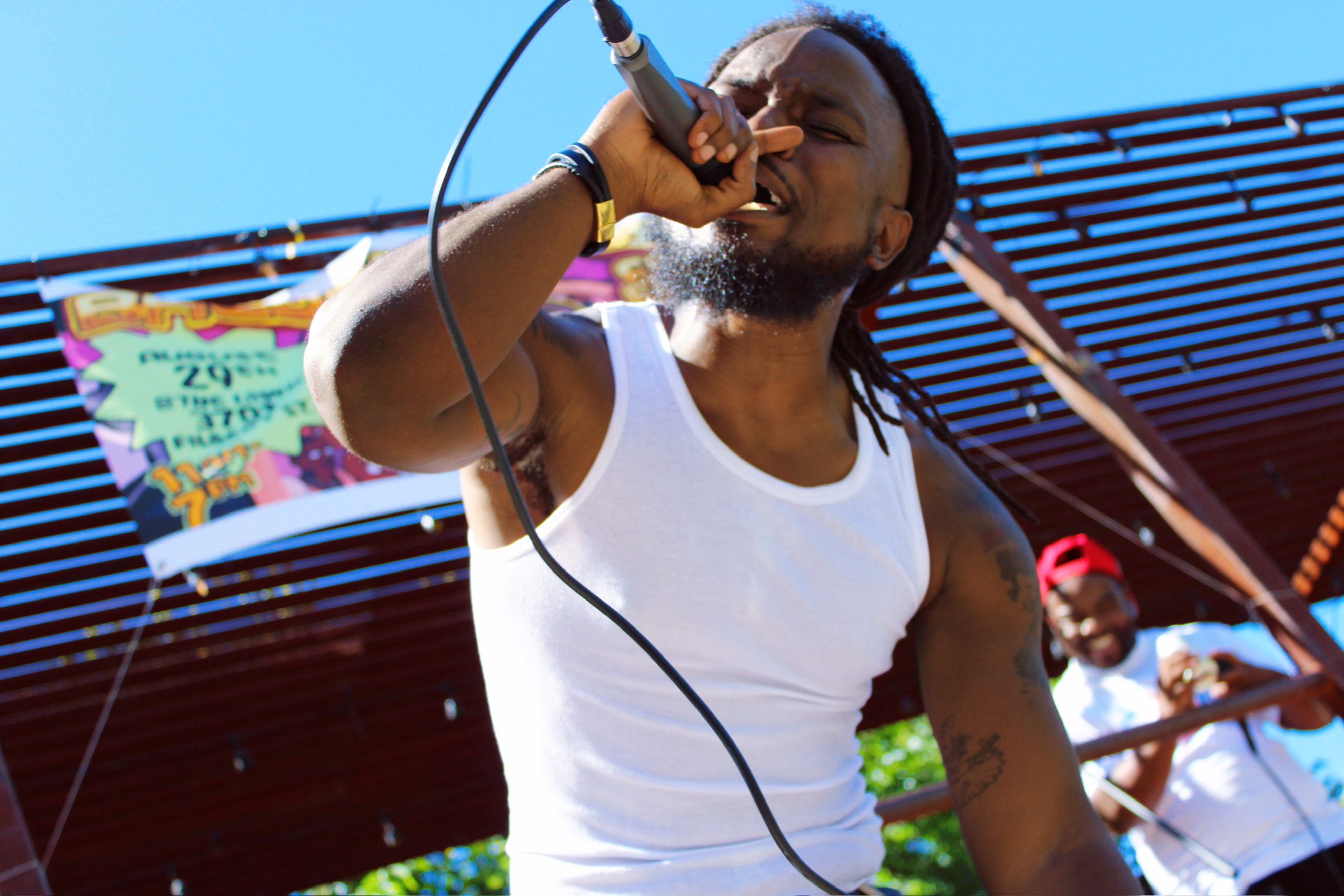 Person singing into a microphone outdoors, wearing a white tank top, with a colorful banner in the background.