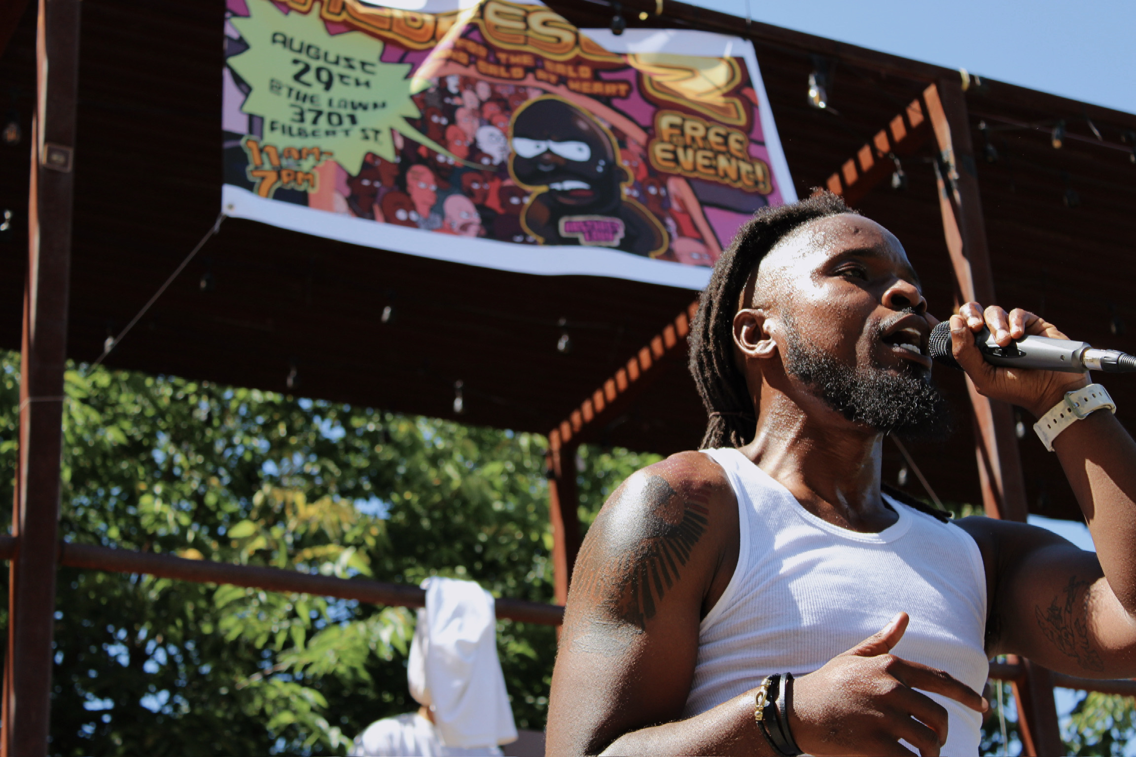 Man with dreadlocks and a beard wearing a white tank top, speaking into a microphone outdoors under a stage, with trees and a billboard in the background.