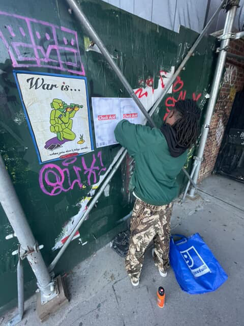 Child in green jacket and camouflage pants spray-paints graffiti on a green wall, with a blue bag and sidewalk nearby.