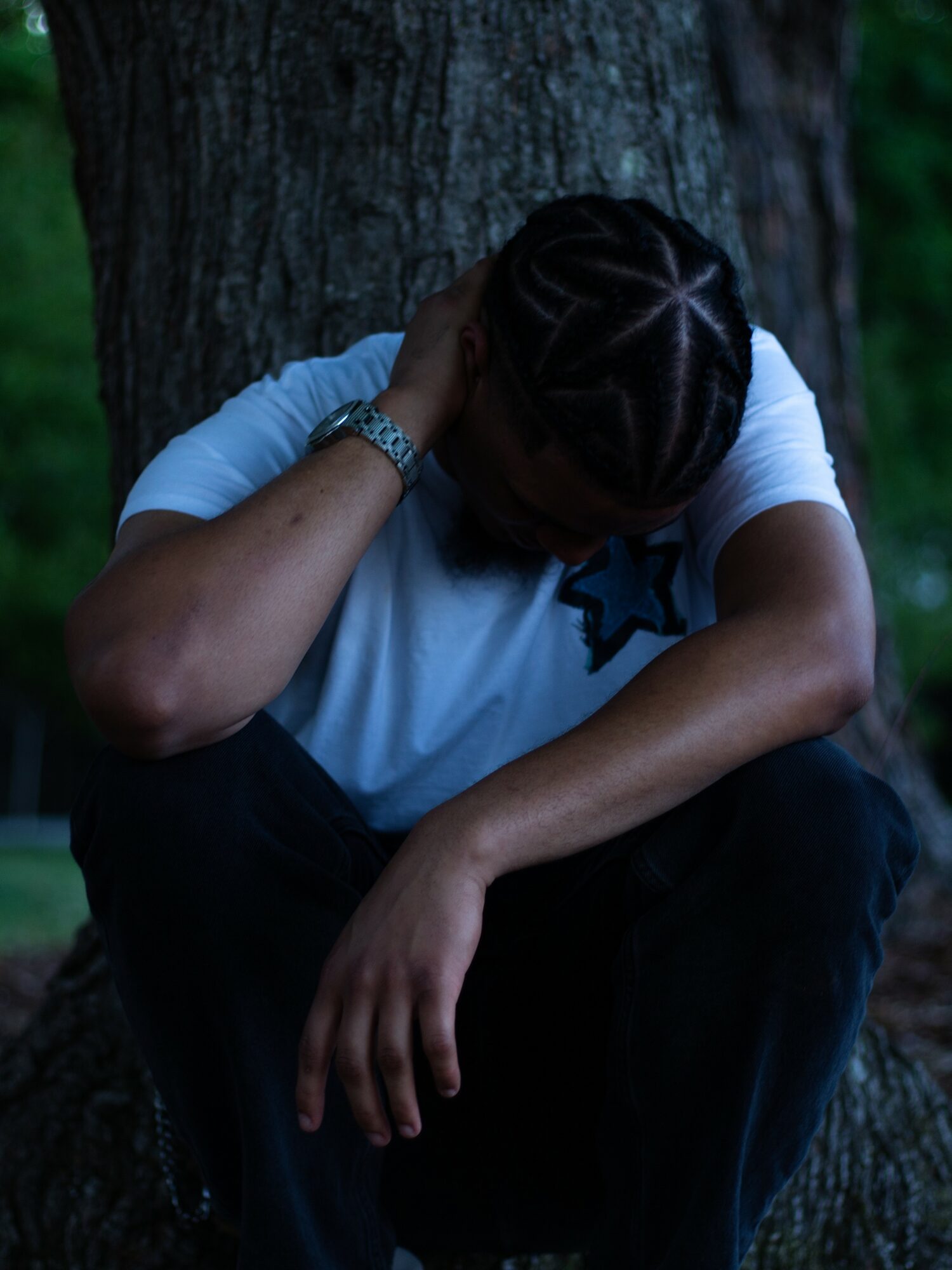Person sitting with head down, arm resting on knee, outdoors near a tree, wearing a white shirt and watch.